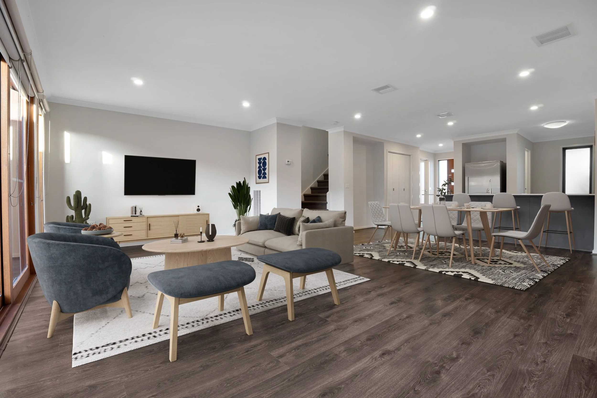 Open-concept living and dining room with wooden flooring, beige sofa, blue chairs, flat-screen TV, tall green plants, and a round coffee table in the living area; stylish dining table with six chairs near the kitchen with stainless steel refrigerator.