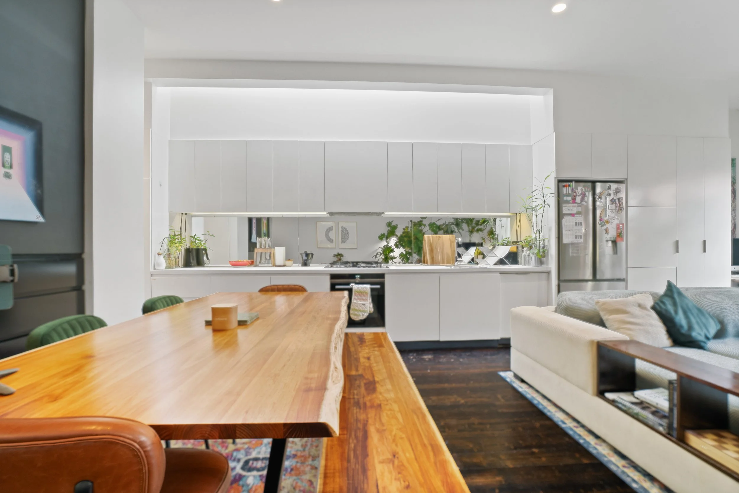 Open-concept living and kitchen area with white cabinets, a large wooden dining table, and a beige sofa with cushions. Decor includes plants and wall art.