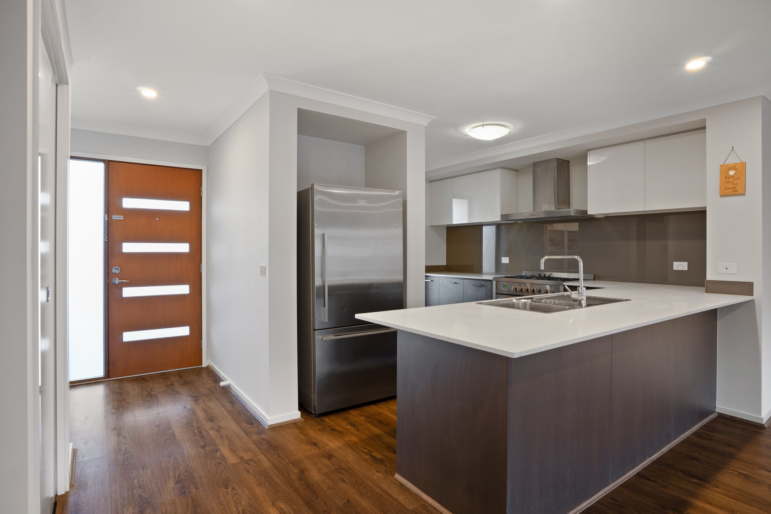Modern kitchen with wooden front door, stainless steel refrigerator, stove, beige countertops, dark wood flooring, and light-colored cabinets.