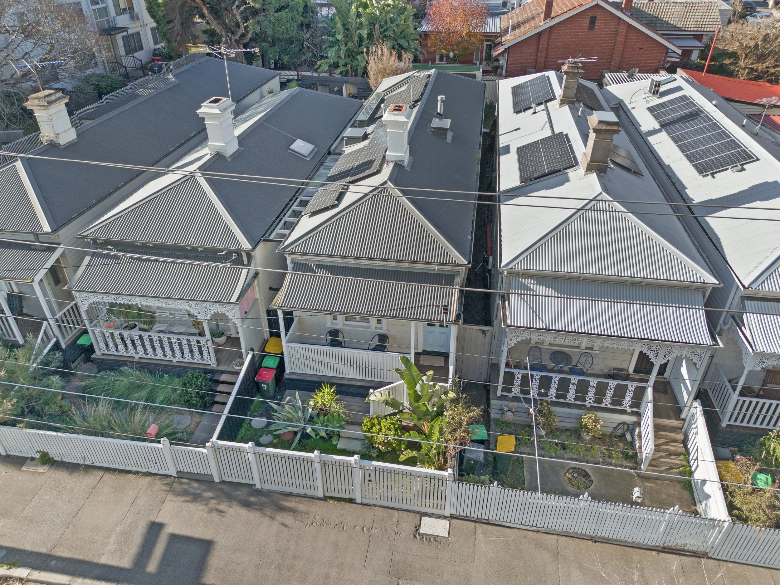 Aerial view of a row of three Victorian-style houses with metal roofs, some with solar panels, and small fenced gardens in front.