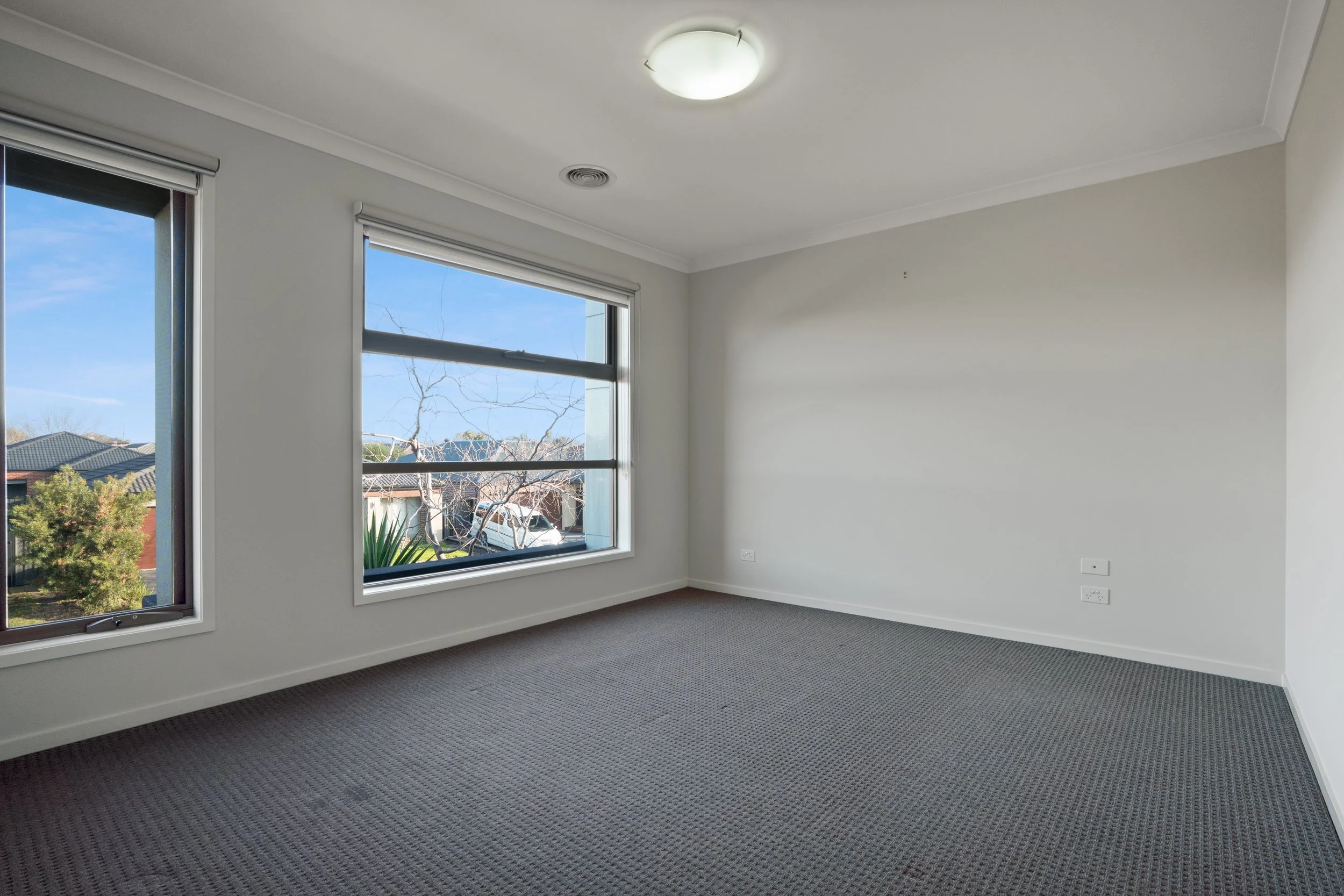 Empty room with beige carpet, white walls, two large windows showing trees and neighboring houses, and a ceiling light fixture.