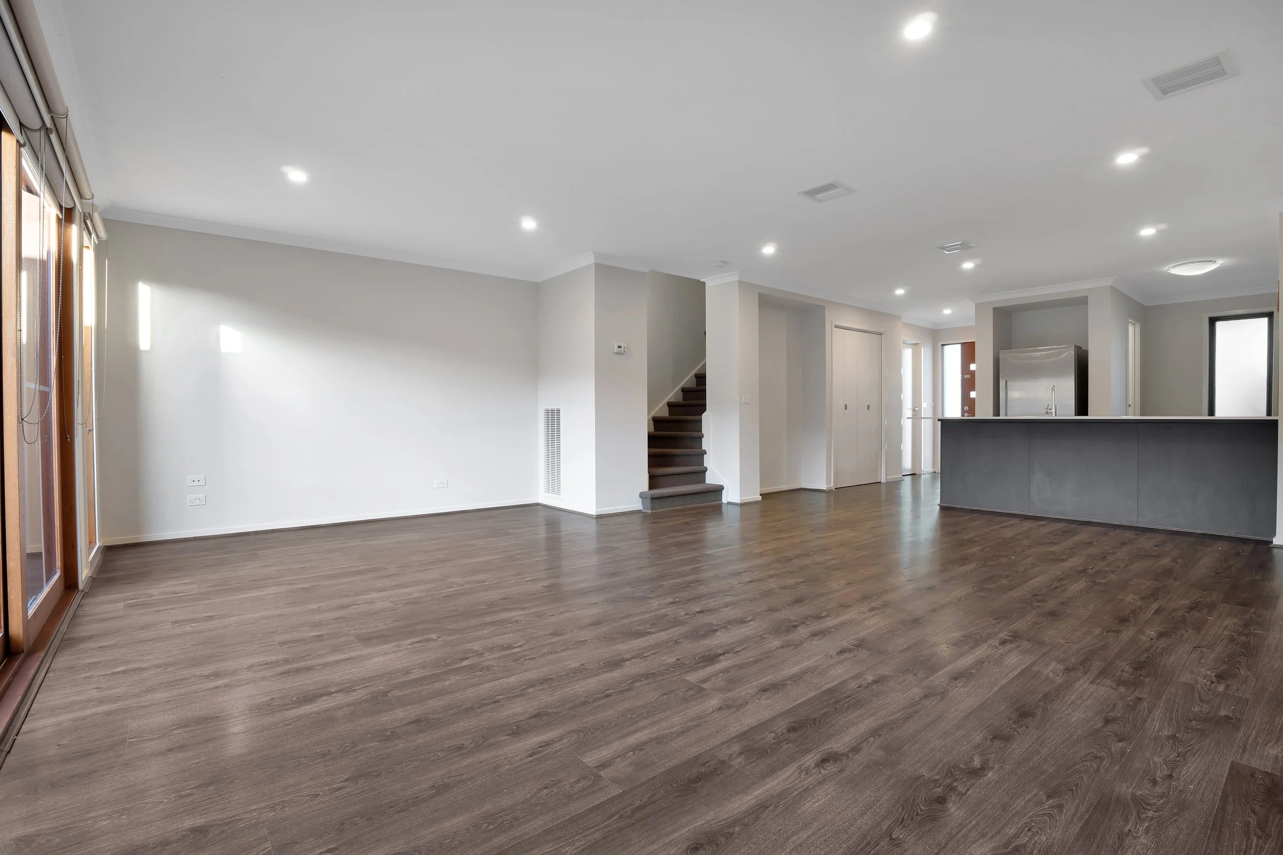 Empty living room with hardwood floors, white walls, and recessed lighting, connected to a kitchen area with a gray island and stainless steel refrigerator.