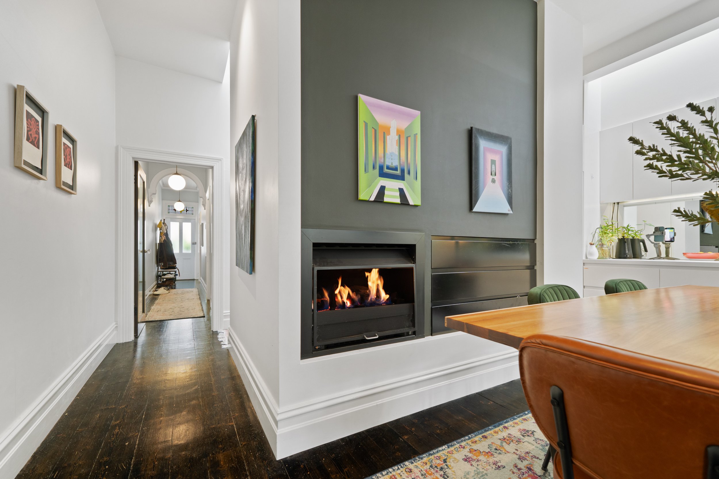 Modern living room with a wall-mounted fireplace, colorful abstract paintings, and a hallway leading to an entryway with a rug and coat rack.
