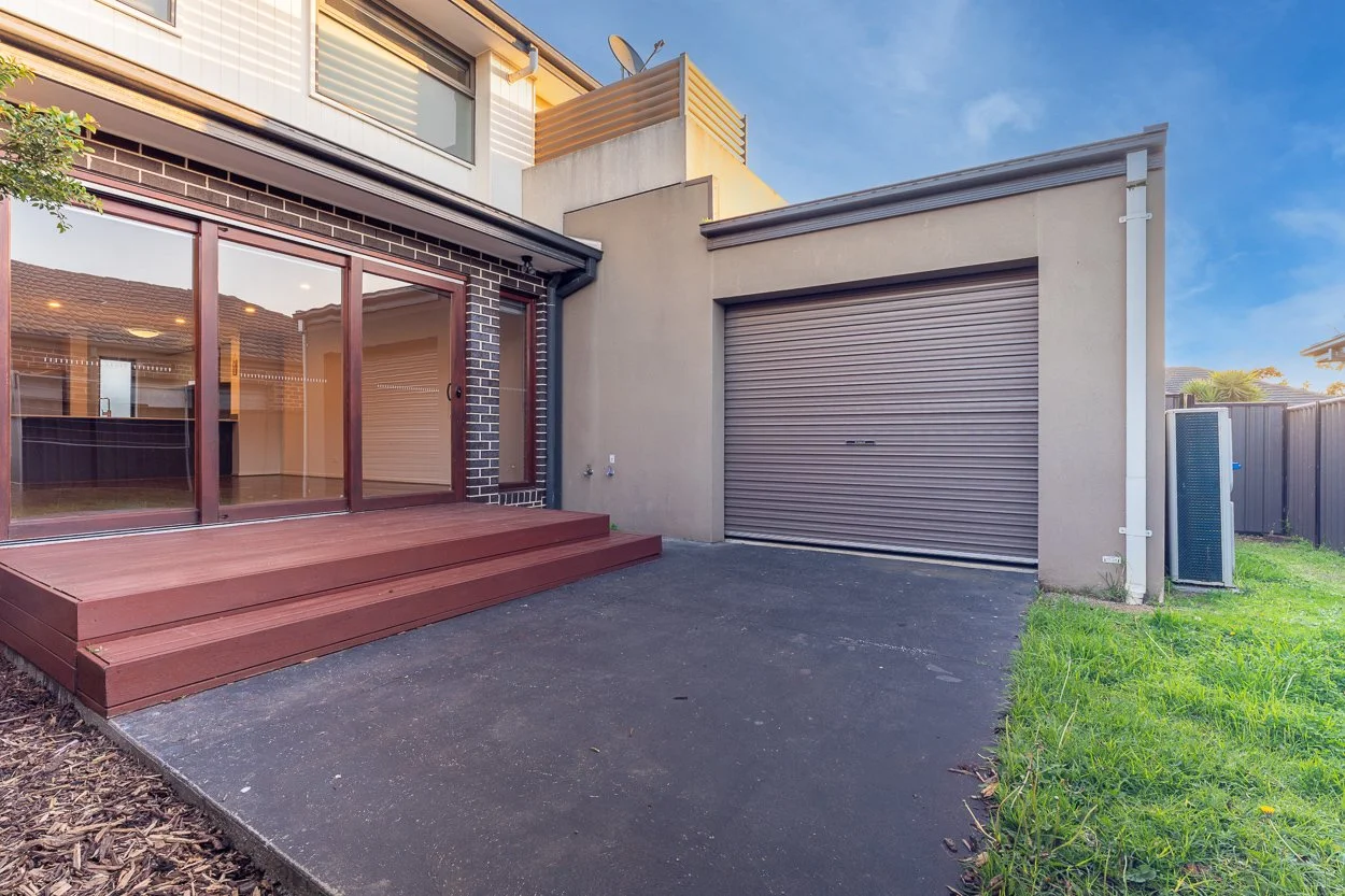Backyard of a modern house showing sliding glass doors, outdoor wooden steps, a closed metal garage door, and a small grassy area. The house has a mix of brick and concrete walls under a blue sky.
