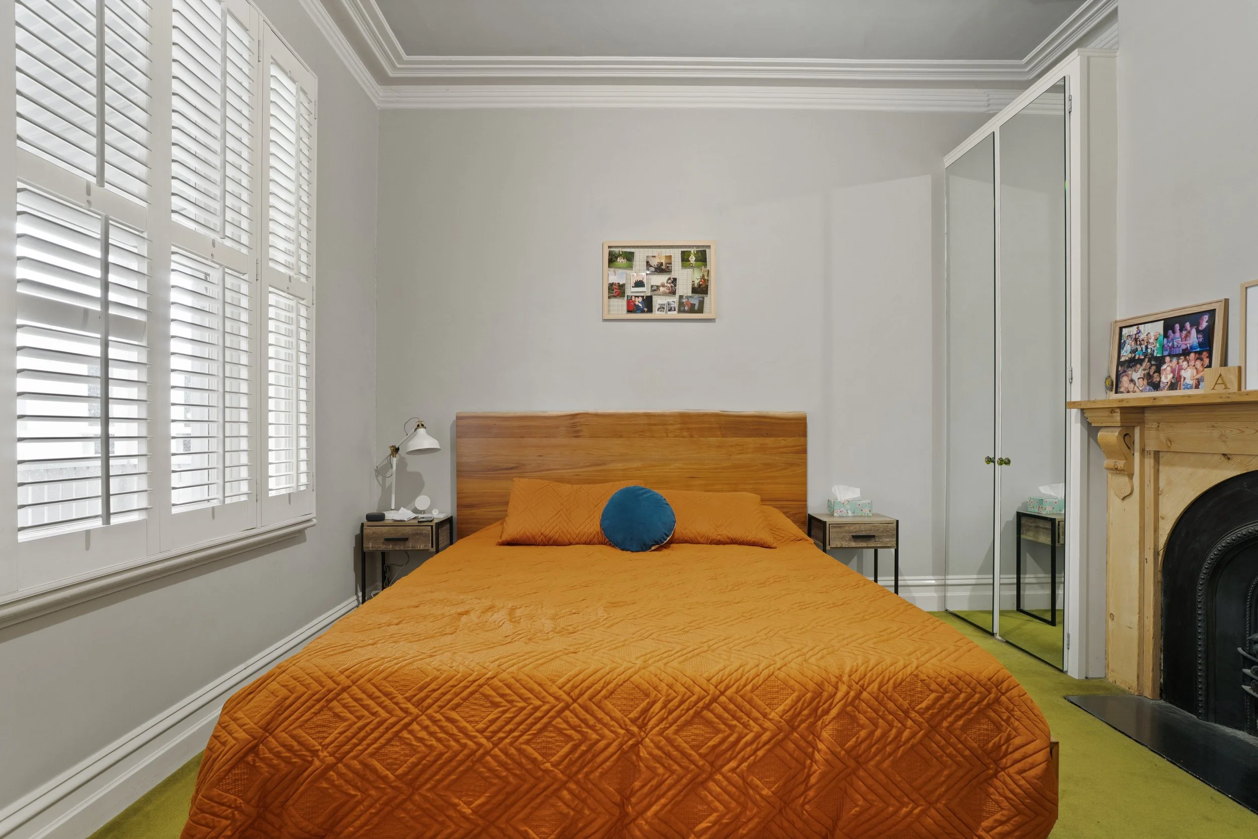 Bedroom with a large bed covered in an orange quilt, a round blue pillow, two bedside tables, a window with plantation shutters, a small collage picture frame on the wall, and a fireplace with framed photos on the mantel.