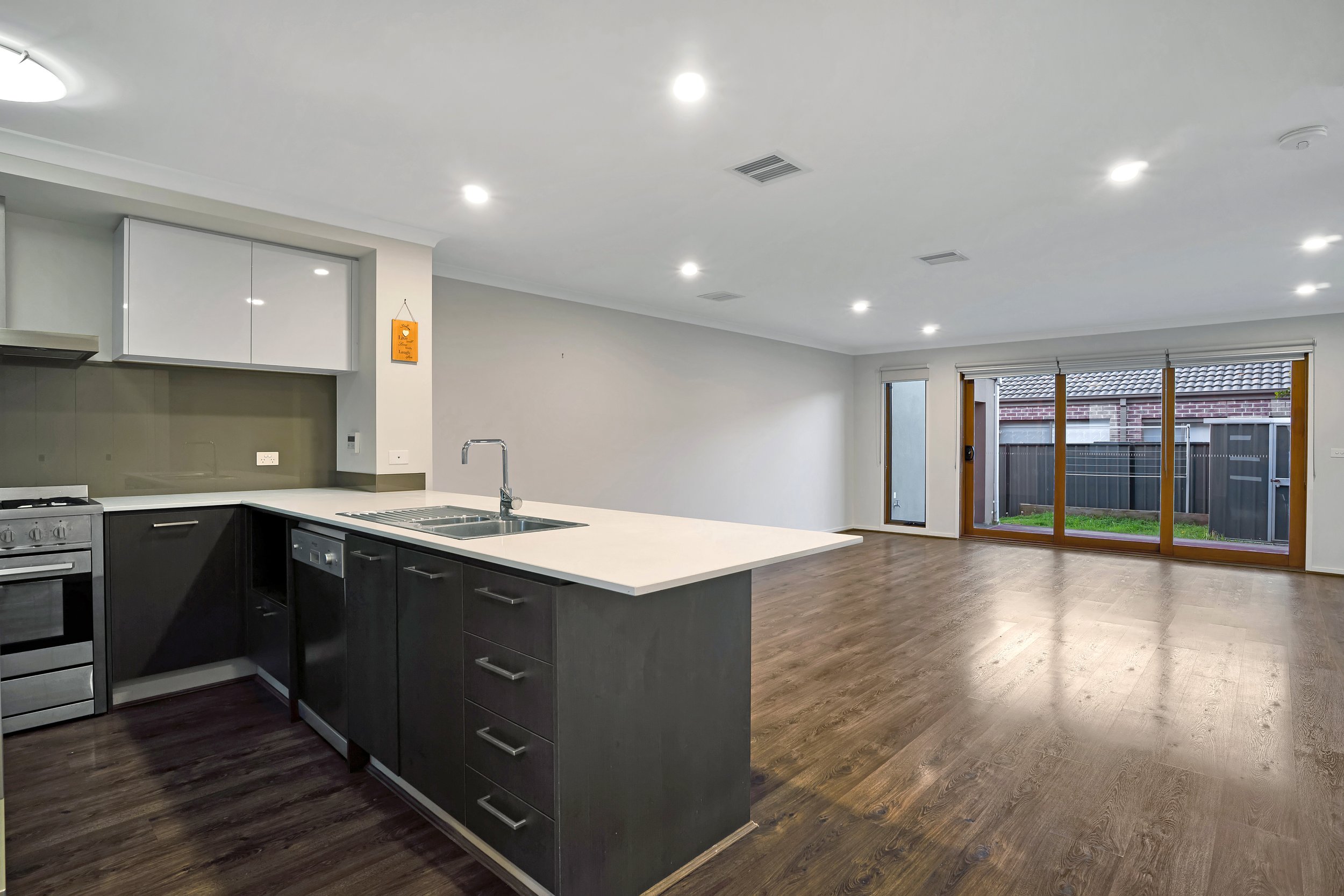 Empty open-concept living area and kitchen with wooden flooring, large glass sliding doors to outdoor yard, white ceiling, and recessed lighting.