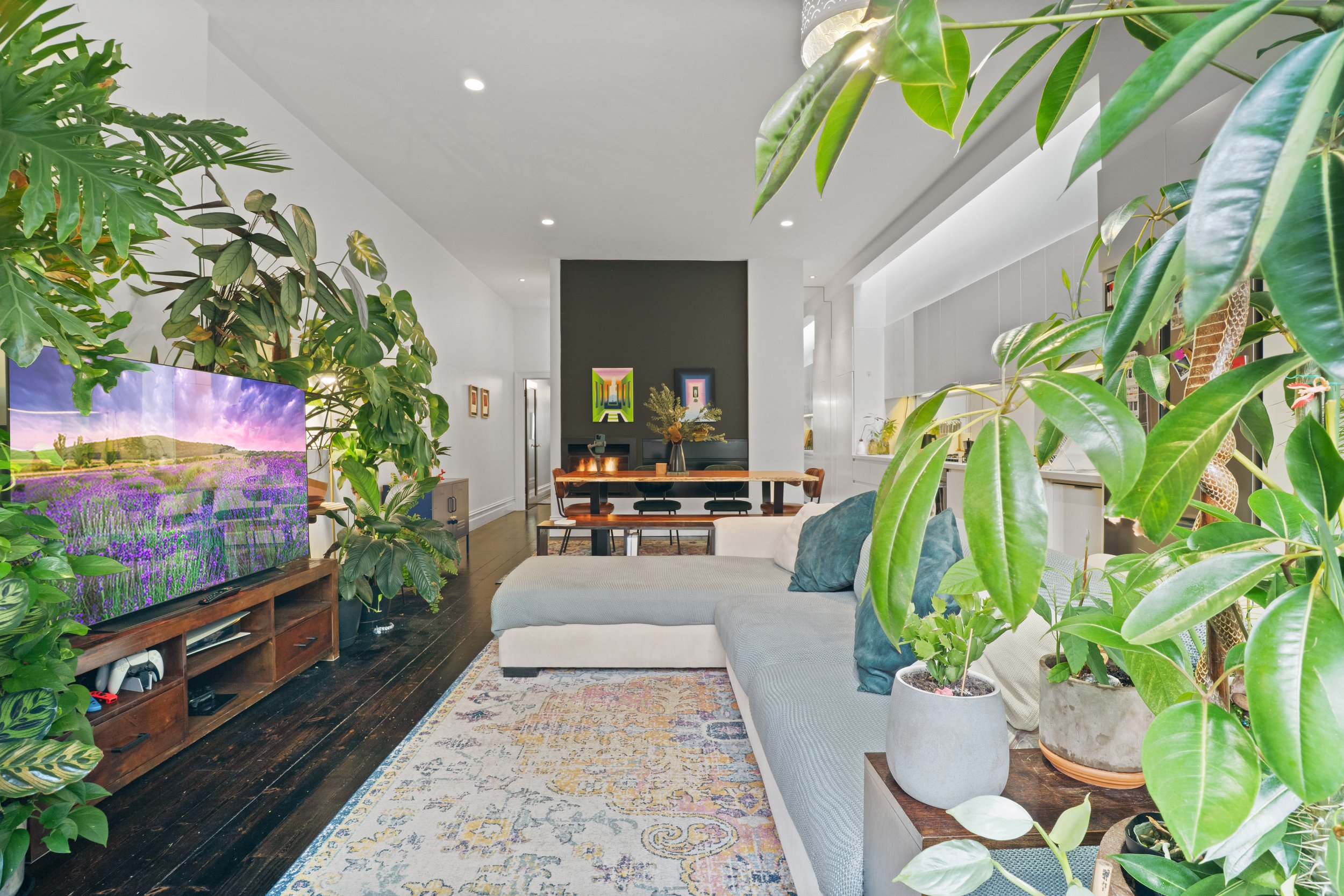 Living room with large green houseplants, a gray sectional sofa, wooden furniture, colorful artwork, and a dark hardwood floor.