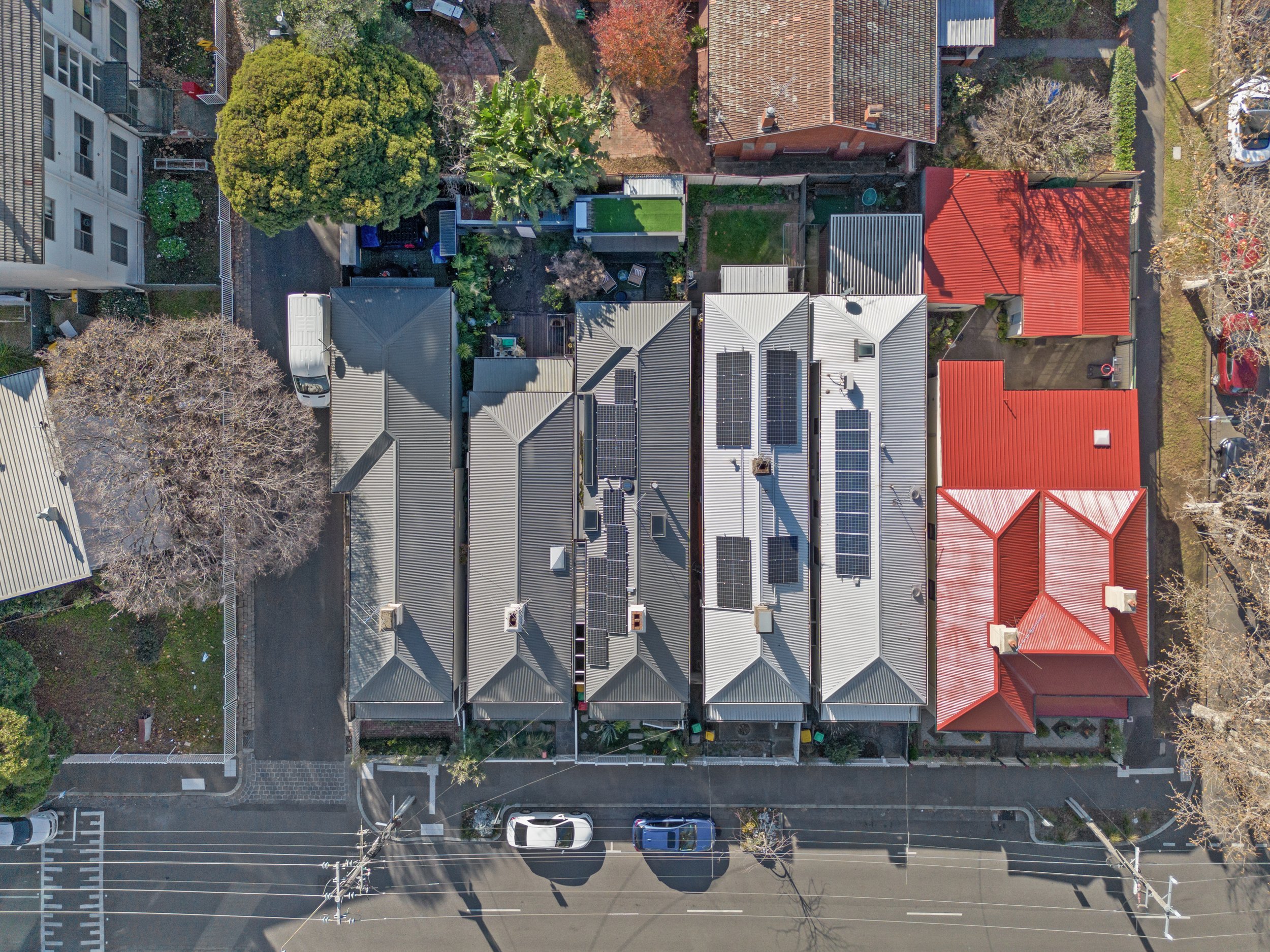 Aerial view of a residential neighborhood with multiple houses, some with solar panels on their roofs, trees, and parked cars along the street.