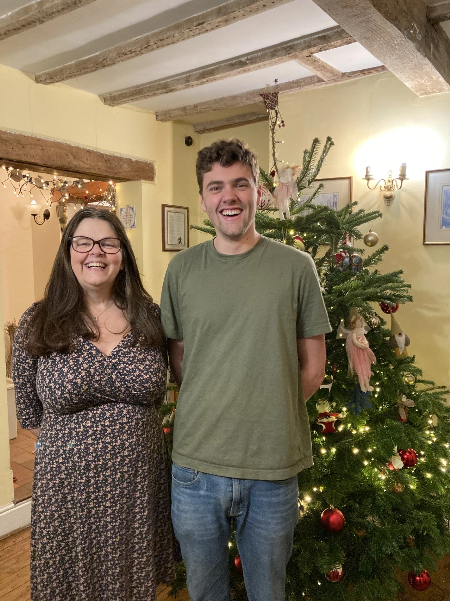 A woman and a young man stand together smiling in front of a decorated Christmas tree, inside a warmly lit room with wooden beams and framed pictures on the wall.