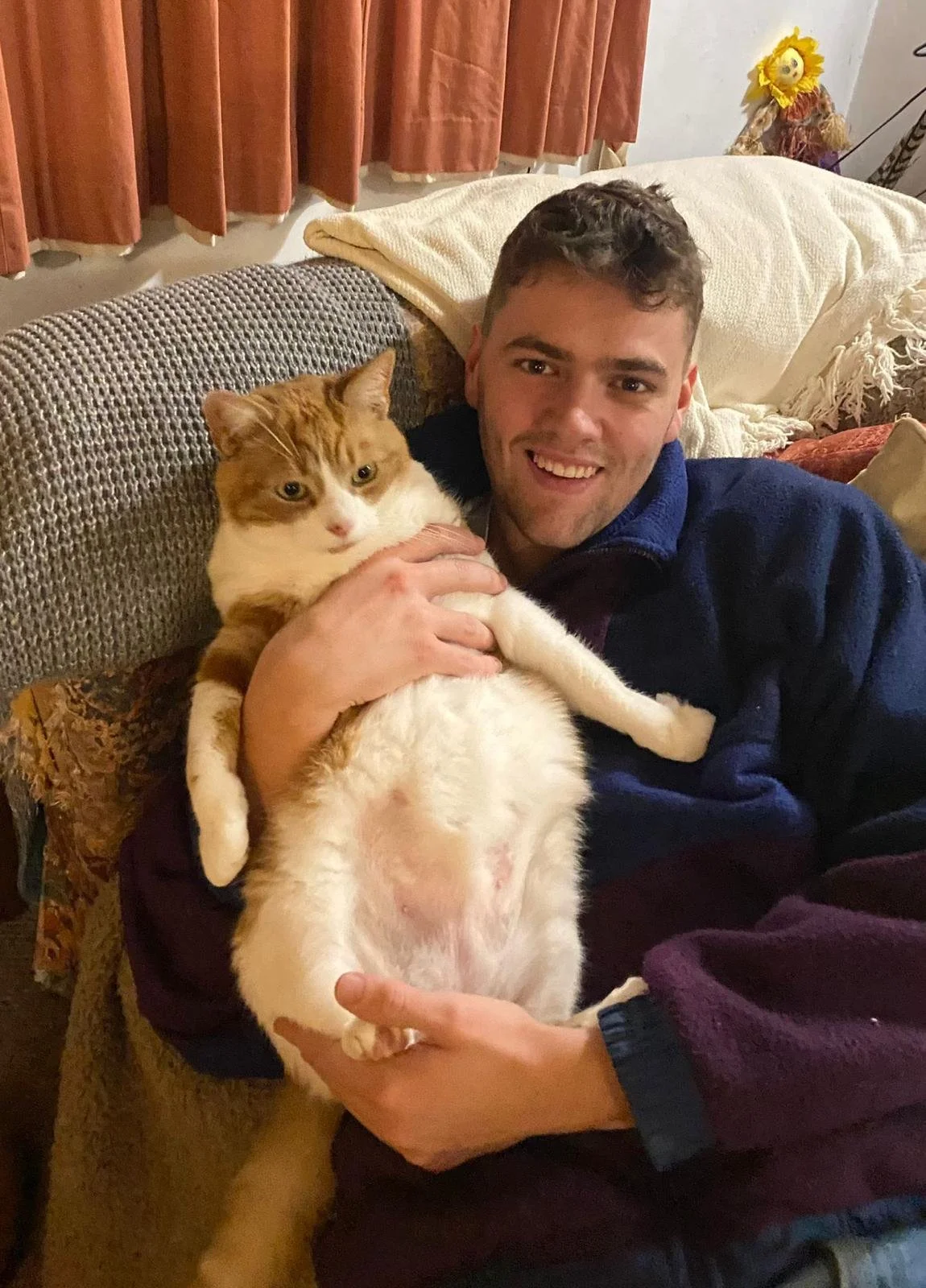 A young man sitting on a beige couch, holding an orange and white cat in his arms, smiling at the camera in a cozy living room with curtains, pillows, and a decorative sunflower in the background.