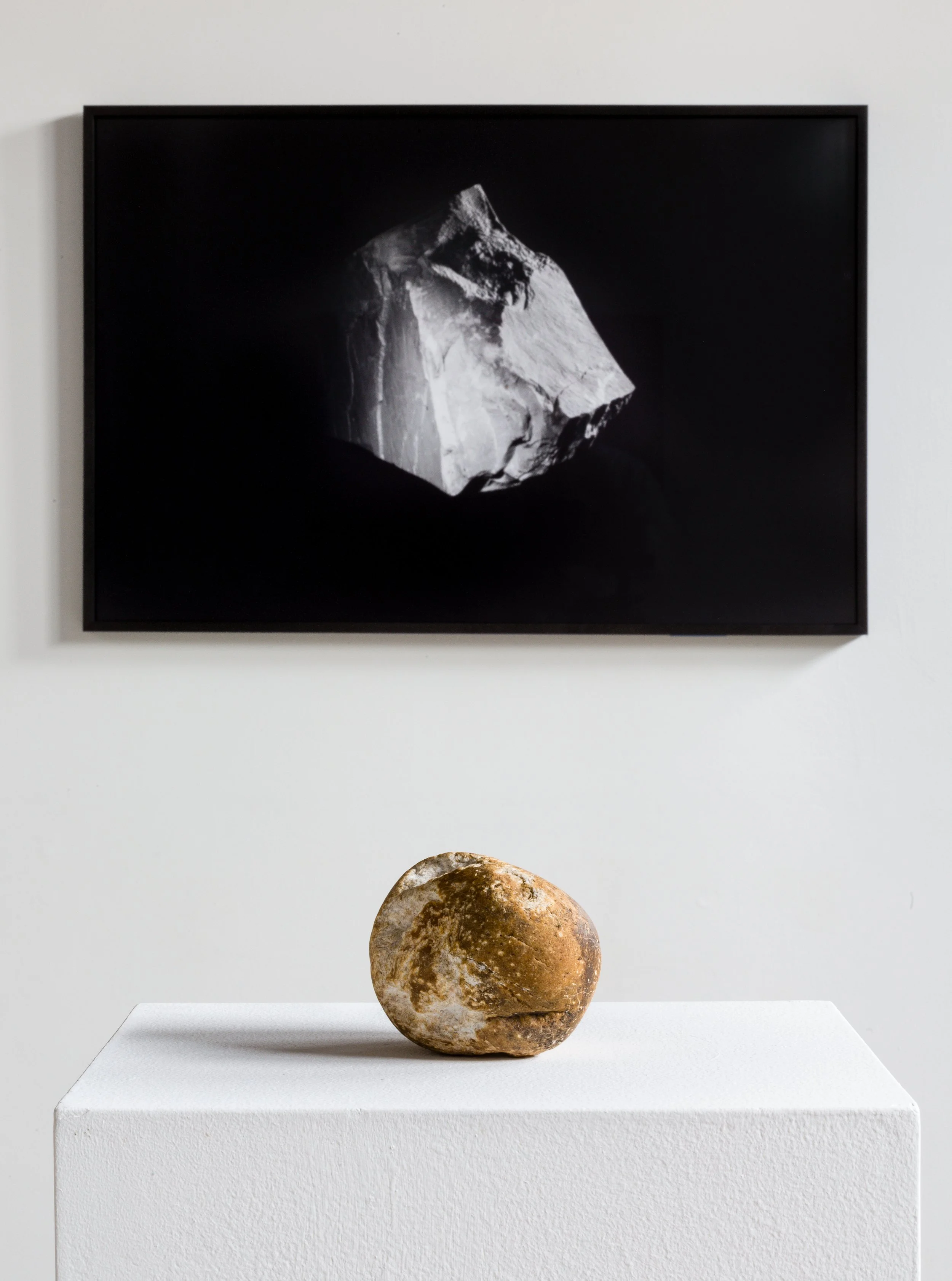 An close-up view of artist Leah Raintree's work featuring a framed black and greyscale 2-d work on a white brick wall and one stone sculpture on a pedestal in the foreground.