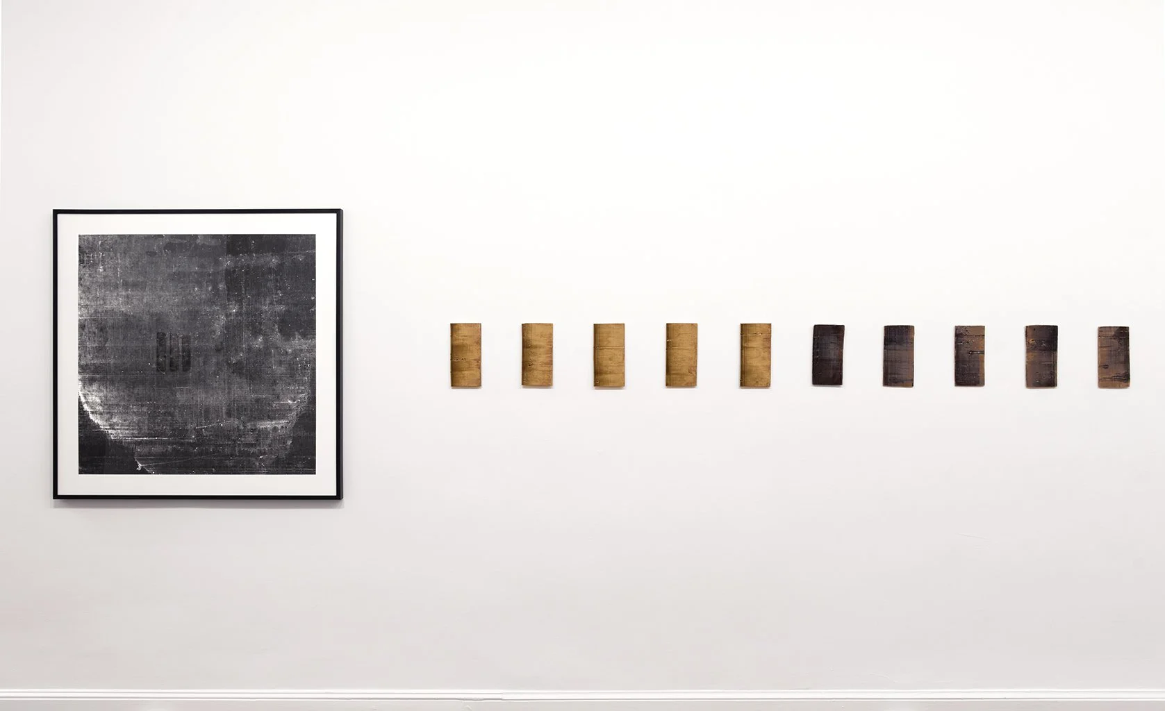 A close up view of multimedia artist Leah Raintree's work featuring one framed dark 2-d work and ten ceramic panels shading from orange to black on a white wall.