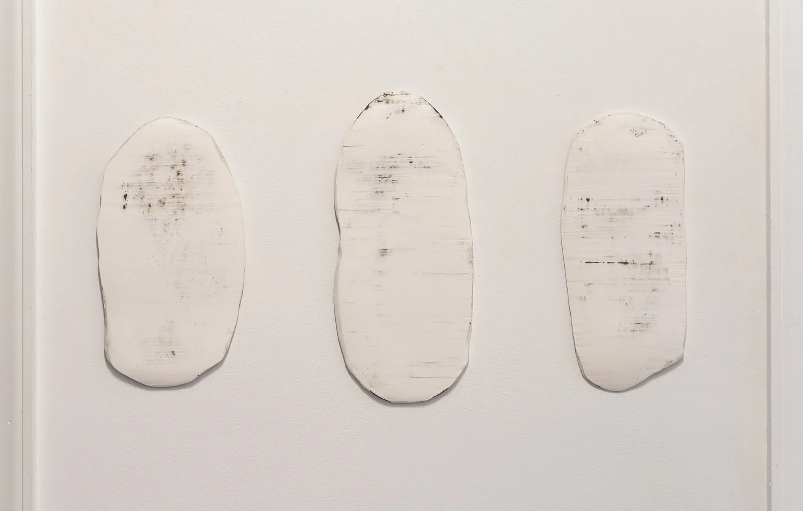 A close up view of Brooklyn based artist Leah Raintree's work featuring three clay oval-shaped textured pieces in shades of white and grey on a white wall.