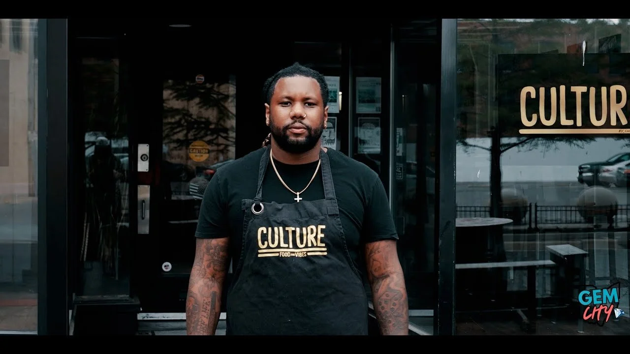 Man standing in front of a restaurant or cafe wearing a black apron with 'CULTURE FOOD VIBES' written on it, black t-shirt, gold necklace, and tattoos on his arms.