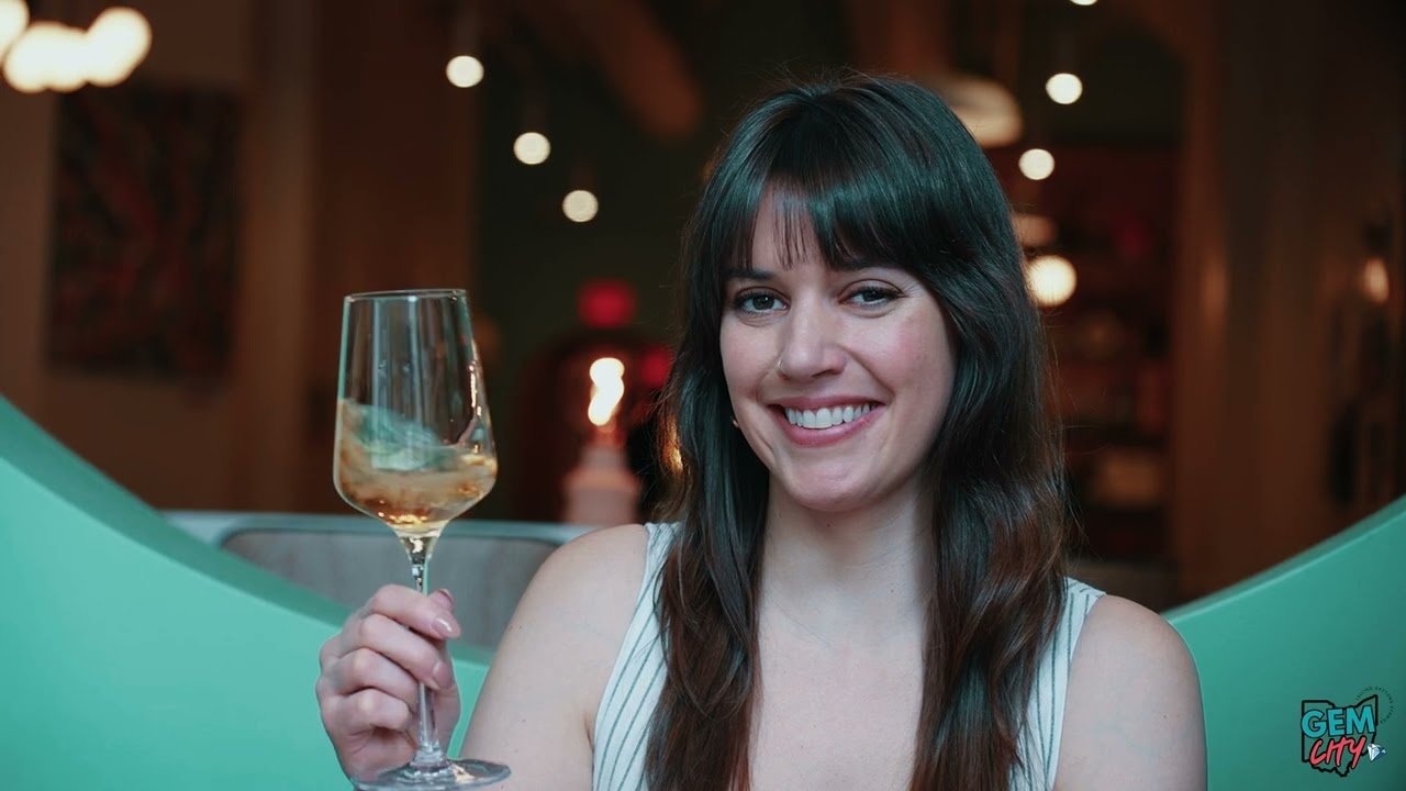 A woman with dark brown hair and a fringe, smiling and holding a glass of white wine in a dimly lit restaurant or bar.
