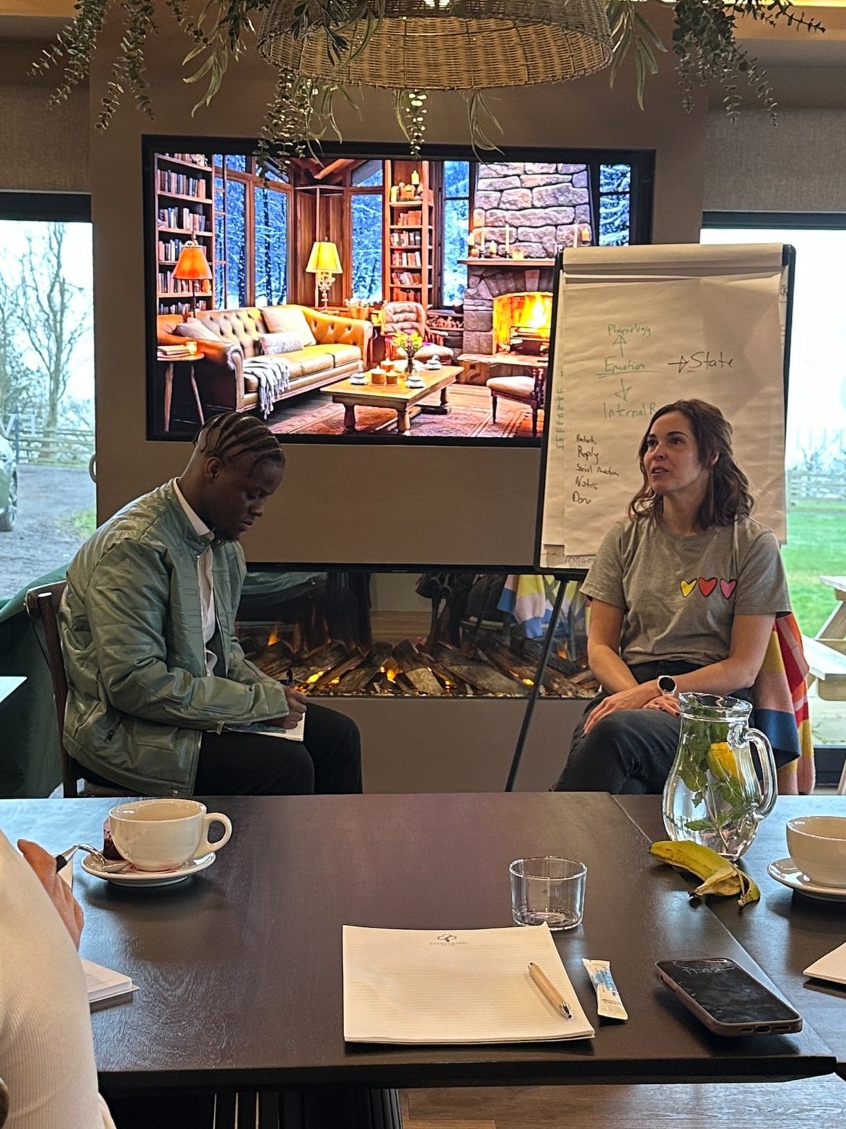 Two people sitting in front of a digital screen displaying a cozy interior with a fireplace. A flip chart with writing is next to them. A table with notepads, a smartphone, cups, a glass, and a pitcher is in the foreground.