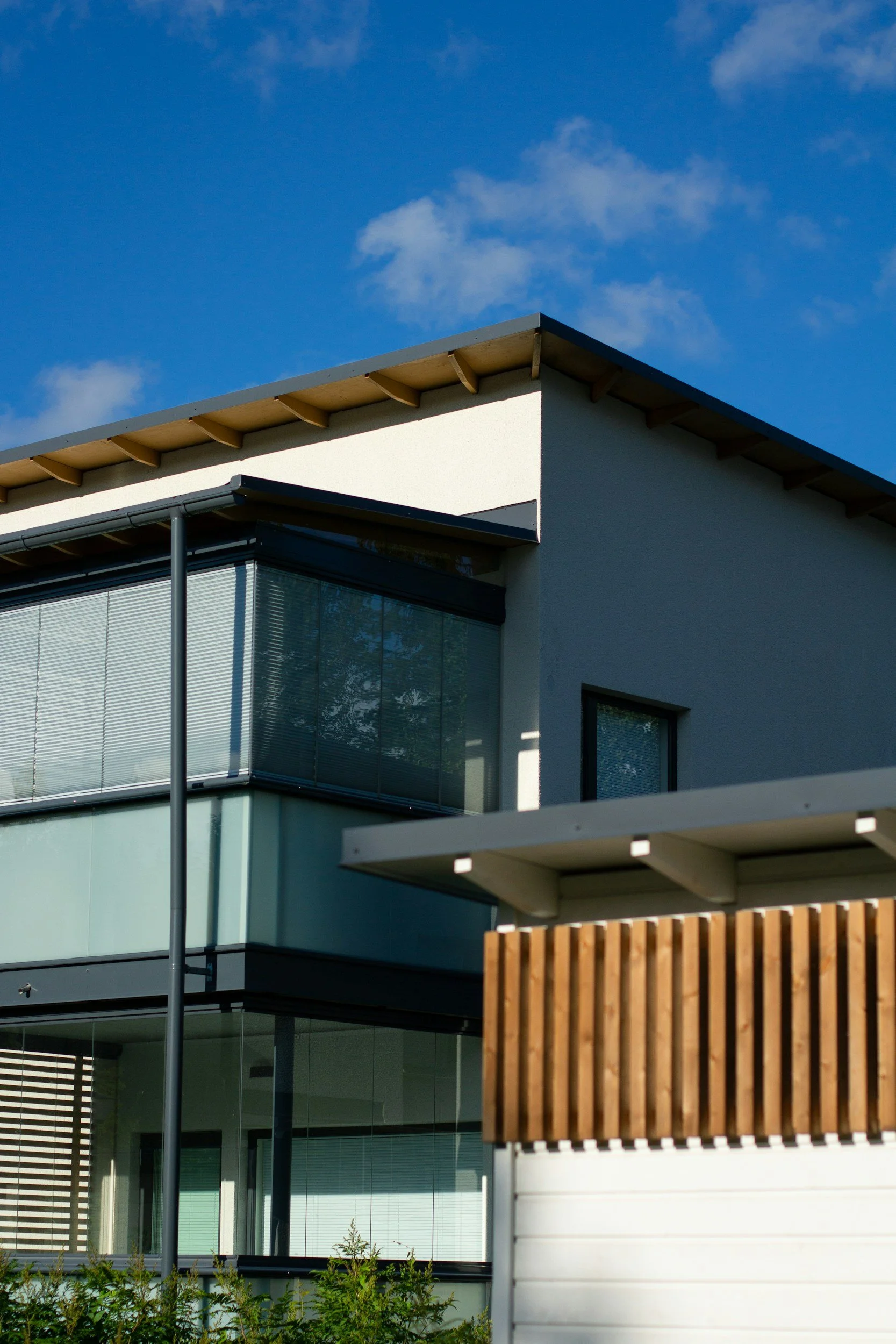 Close-up view of a modern house with large glass windows, white walls, and a sloped roof against a blue sky with a few clouds.
