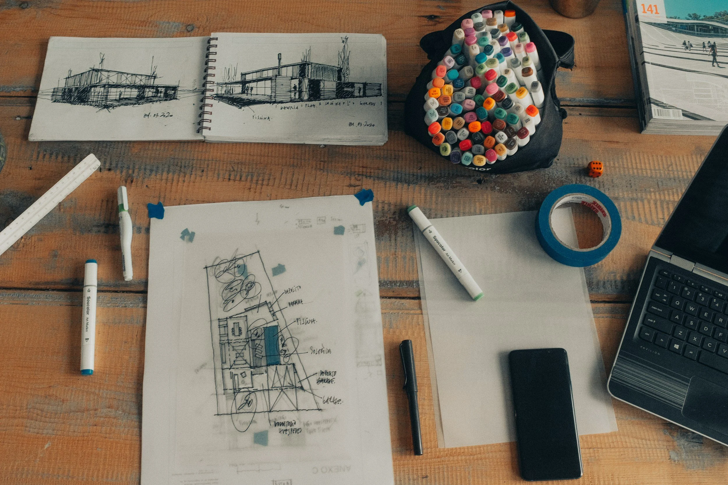 Architectural sketches, markers, washi tape, a smartphone, and an open laptop on a wooden table, indicating a workspace for architectural or design planning.