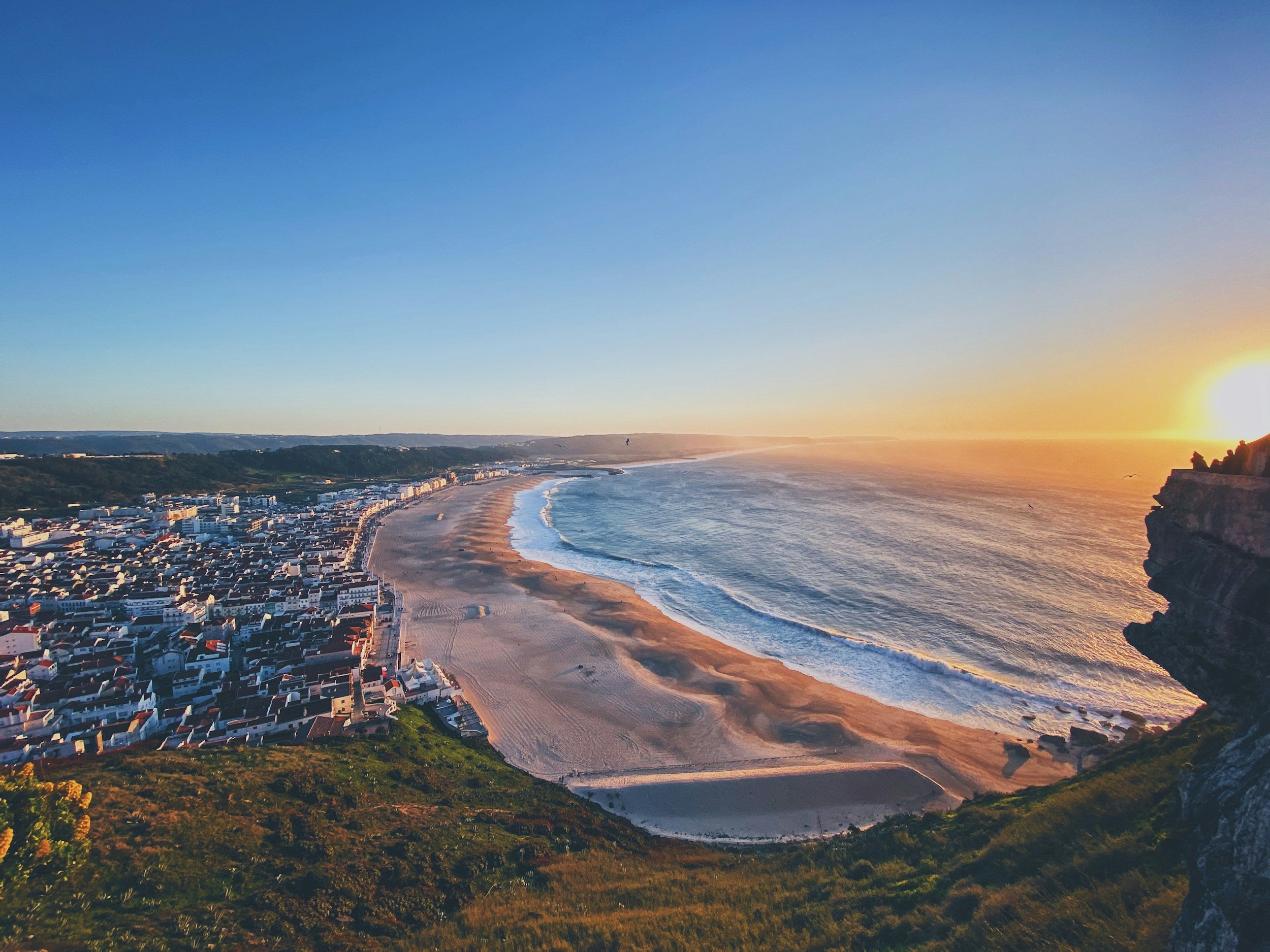 A panoramic view of a coastal town at sunset, featuring a sandy beach, ocean waves, and colorful buildings along the shoreline with a rocky cliff on the right.