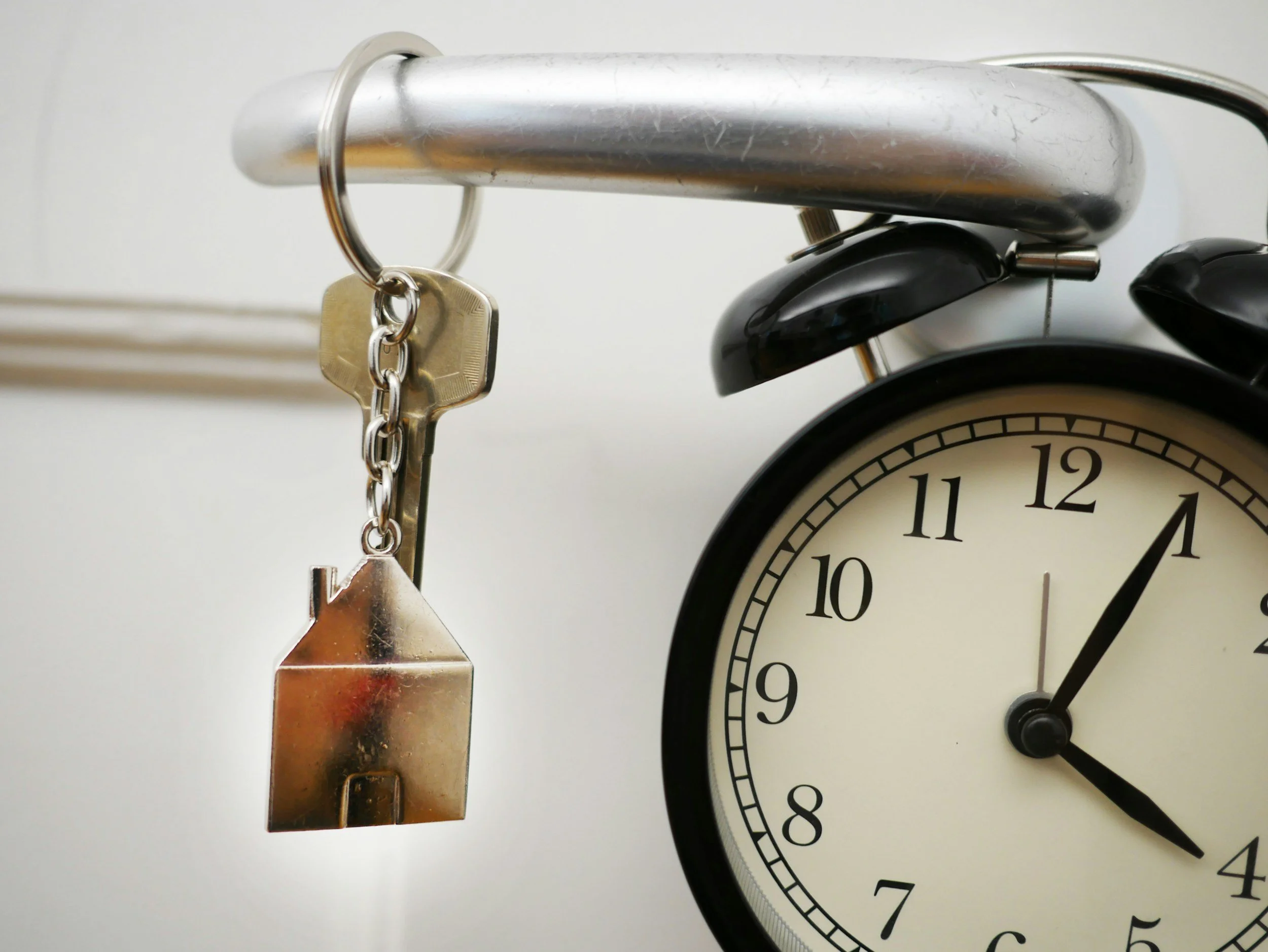 Close-up of a black alarm clock showing 4:05, with keys and a house-shaped keychain hanging from a silver hook above it.