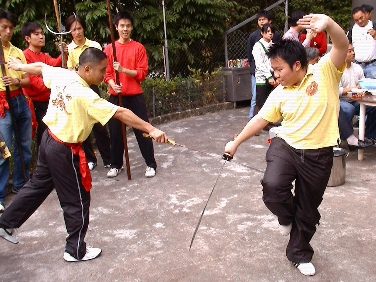 兩位功夫學生參加劍鬥示範，旁觀者在觀看。左側的男子持劍伸出手臂，右側的男子則擺出防守姿勢，劍指著他。背景中有幾位持傘的人，還有一群兒童和成年人觀察這個活動。