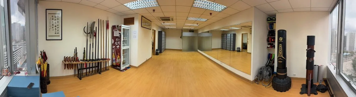 Martial arts training room with wooden floors, large mirror, weapons display, punching bags, and exercise equipment near windows showing cityscape