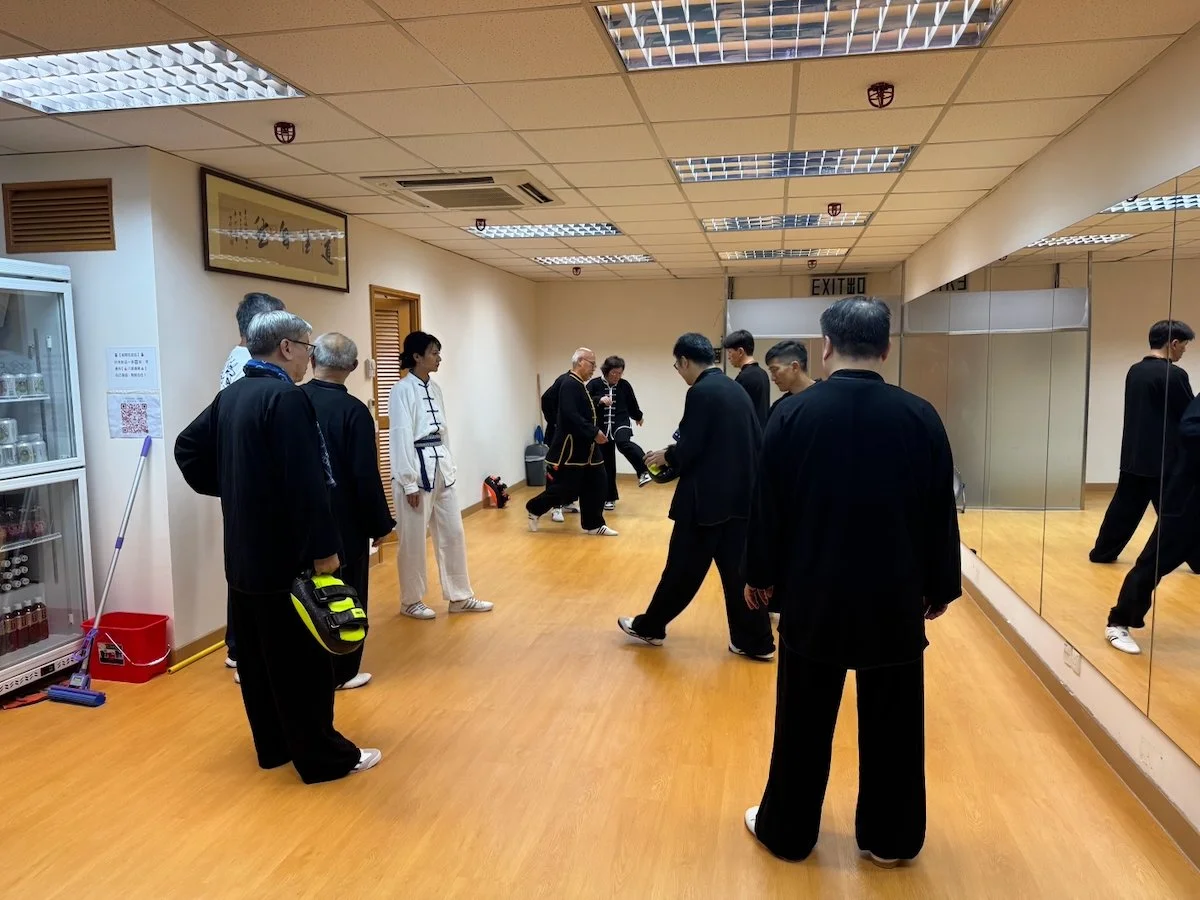 Group of people in a martial arts classroom with wooden floor, mirrored wall, and ceiling lights, some practising moves, others observing.