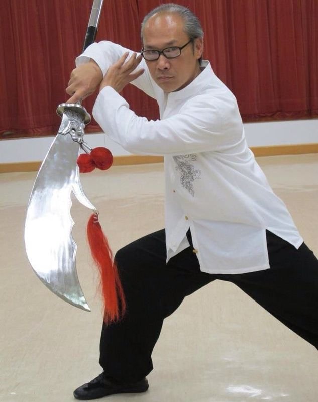 A man dressed in traditional martial arts attire demonstrates a martial arts pose with a large, ornate sword in a room with red curtains.