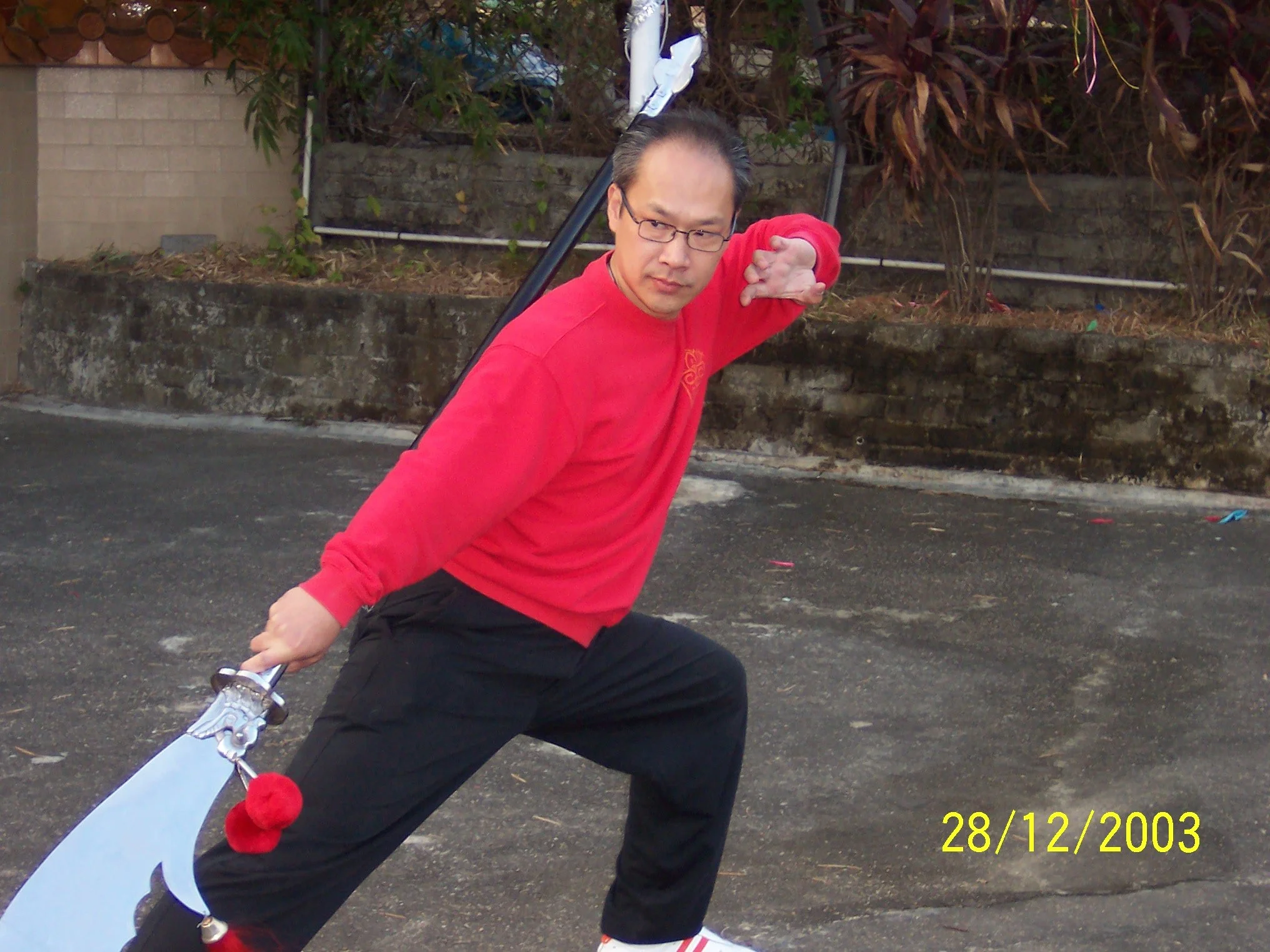 一位穿紅色運動衫和黑色褲子的男子在戶外練習中國武術，持中國武術板劍擺出準備姿勢，背景是磚牆和植物。