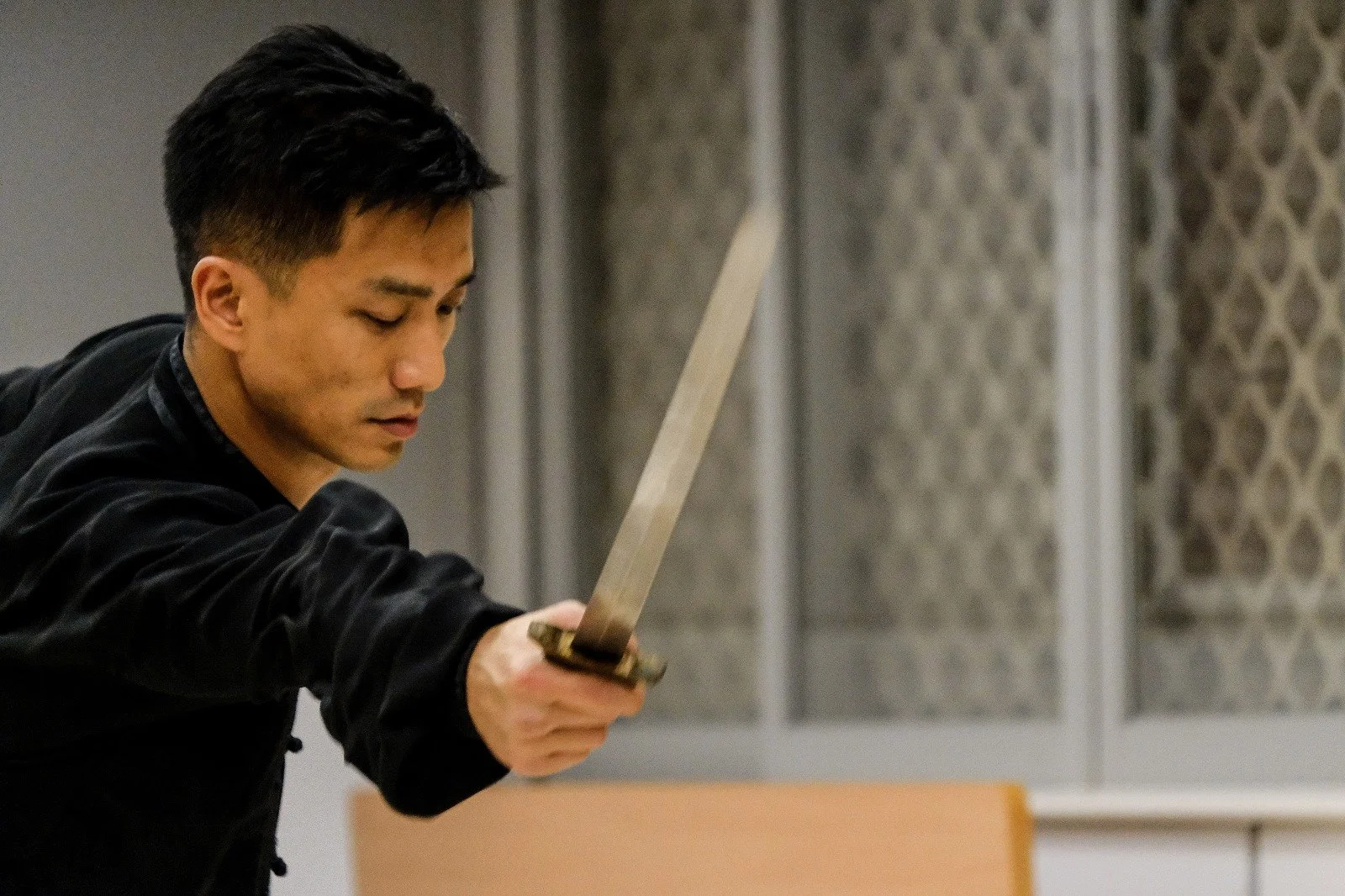 A man is holding a sword in a ready position, with a focused expression, indoors near textured windows.