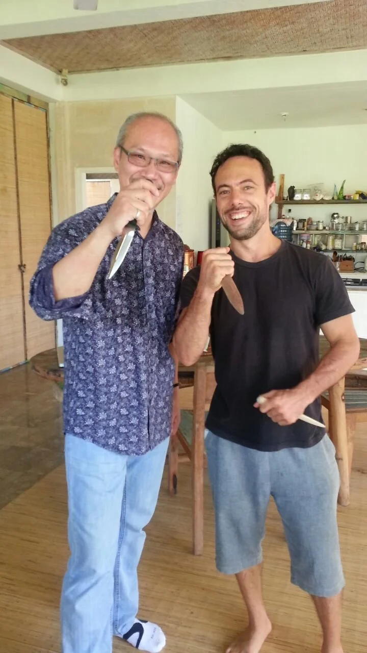 Two men smiling and holding knives in a kitchen.