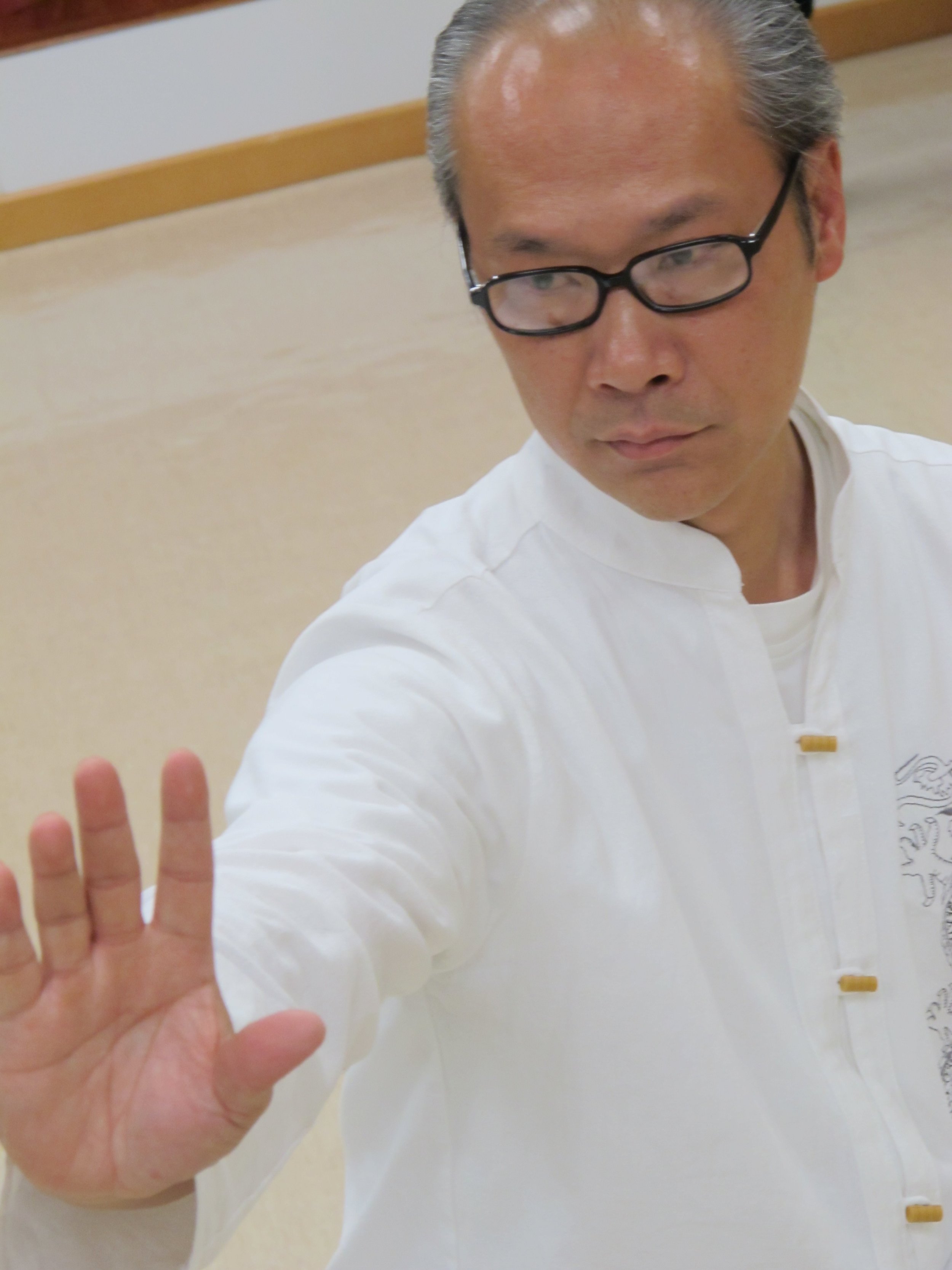 A man wearing glasses and a traditional white martial arts uniform is showing the palm of his hand while looking to the side.