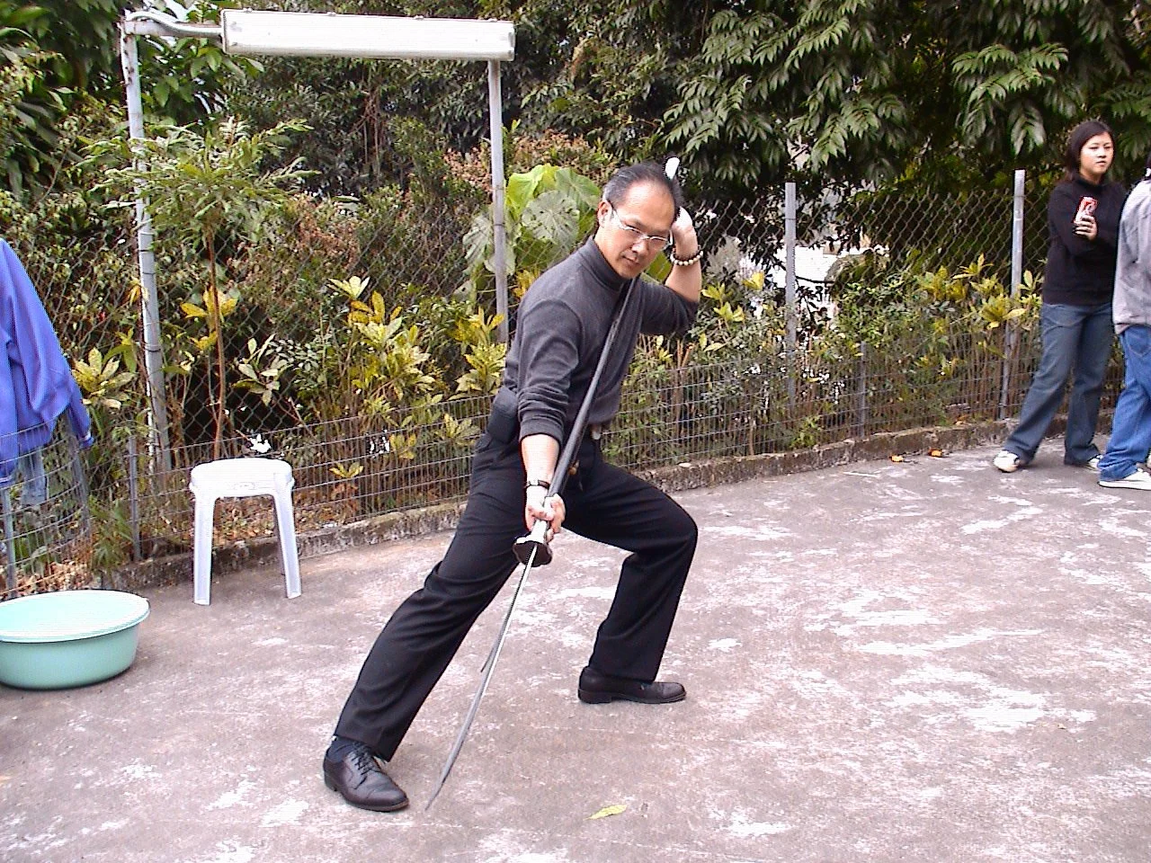 一位穿黑色服裝的男子擺出中國武術姿勢，手持劍，站在戶外，背景有其他人。