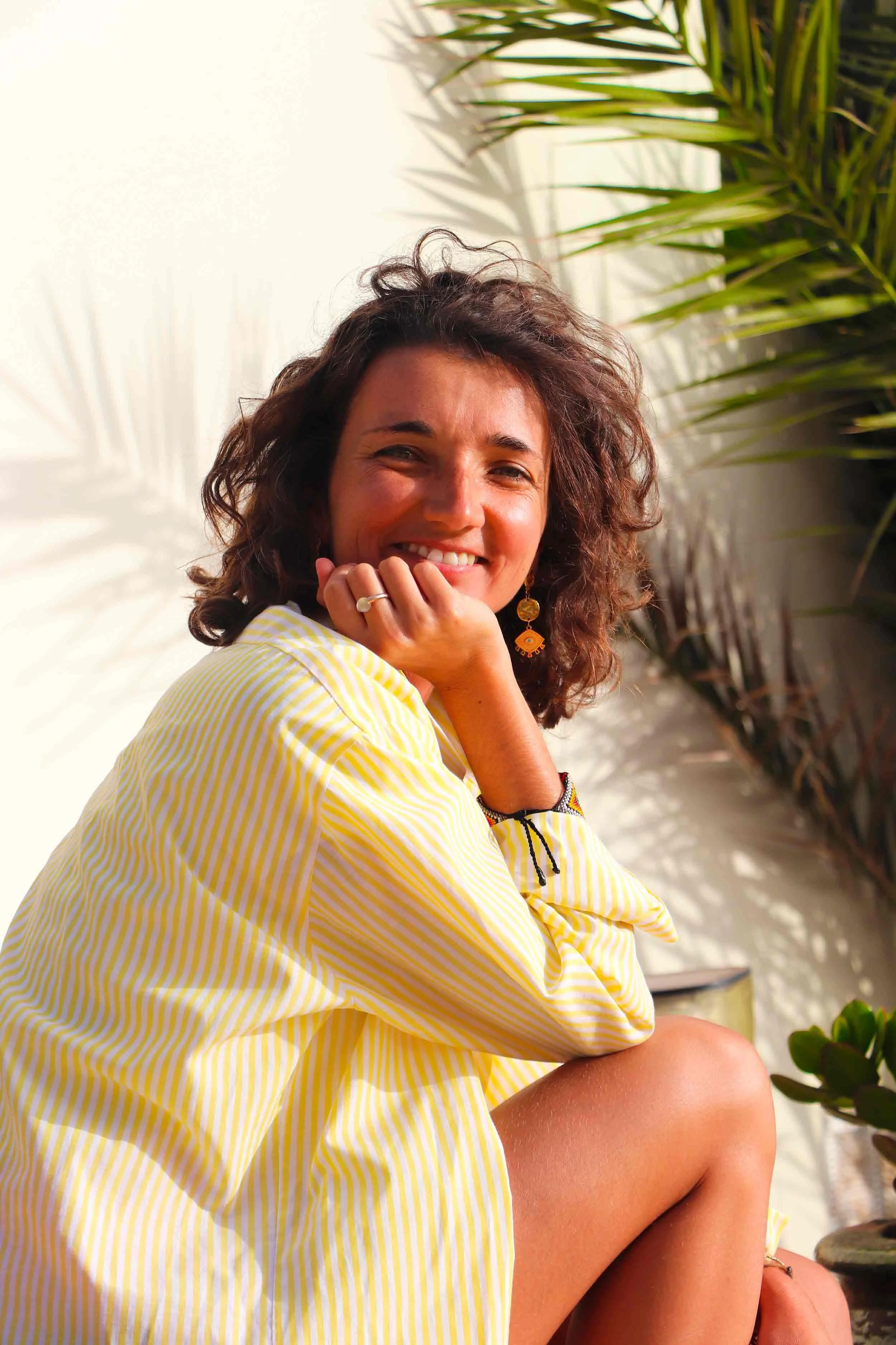 Joséphine, coach business, une femme souriante avec des boucles, portant une chemise jaune à rayures, assise près d'une plante verte, en plein air avec un fond blanc et des ombres de feuilles.