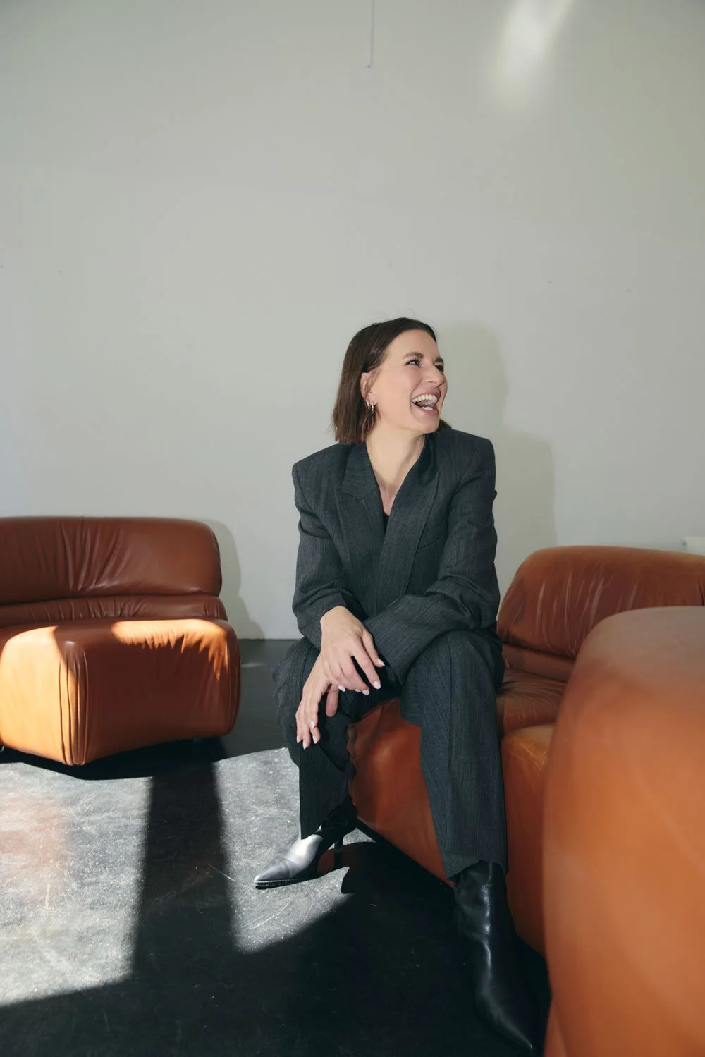 Valerie Junger im grauen Anzug sitzt lachend auf einem braunen Ledersofa – 1:1 Coaching für berufliche Klarheit und Neuorientierung.