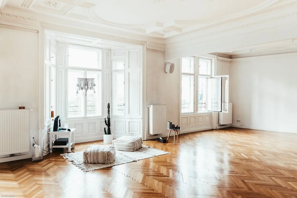 Ein helles, leerstehendes Wohnzimmer mit Holzboden und großen Fenstern, wenig Möbel, einige Kissen, eine Pflanze, eine kleine Tischlampe und eine Stehlampe.