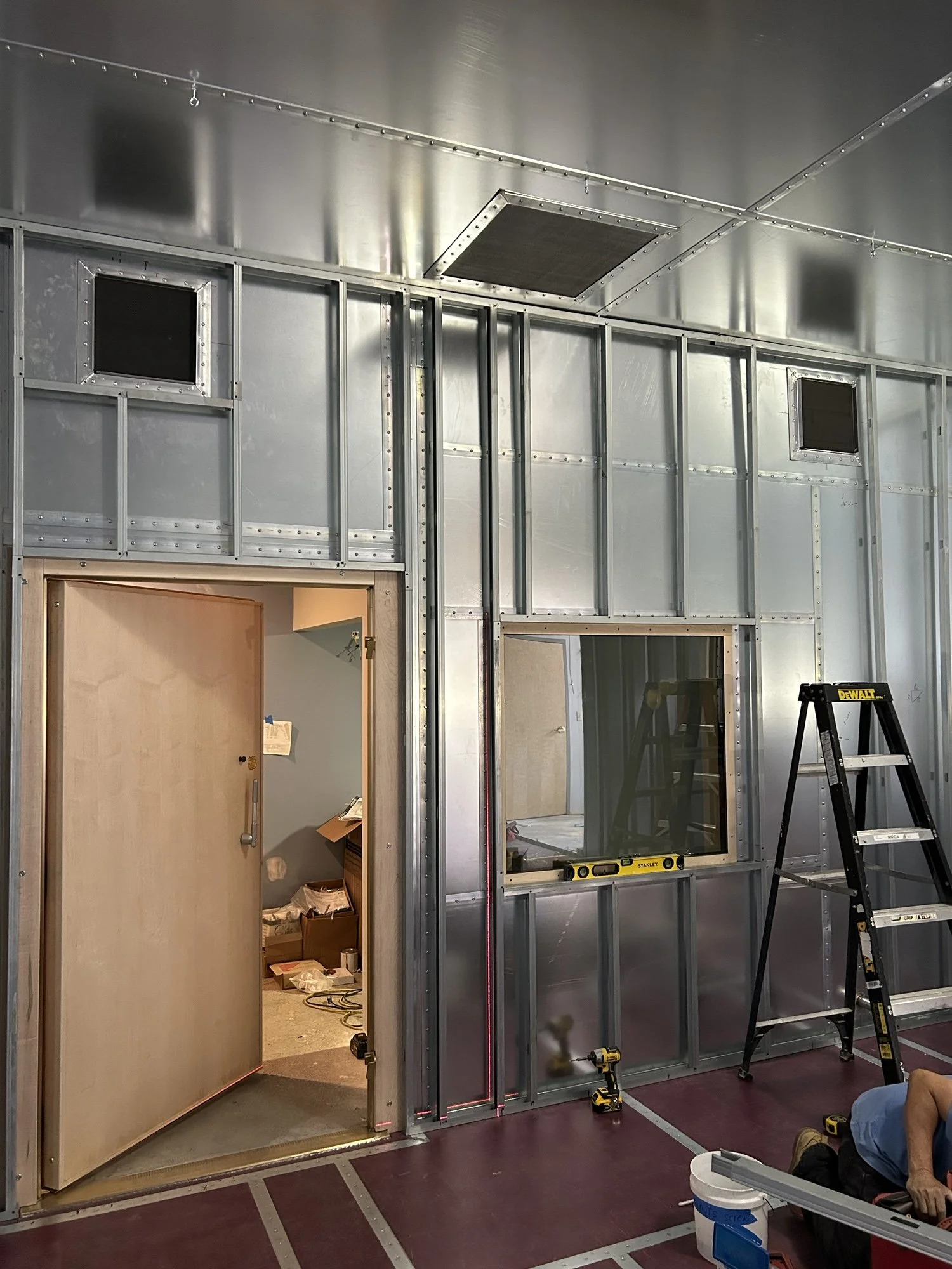Construction site with metal framing and an open door leading into a room. A ladder and tools are present, and a person is working.