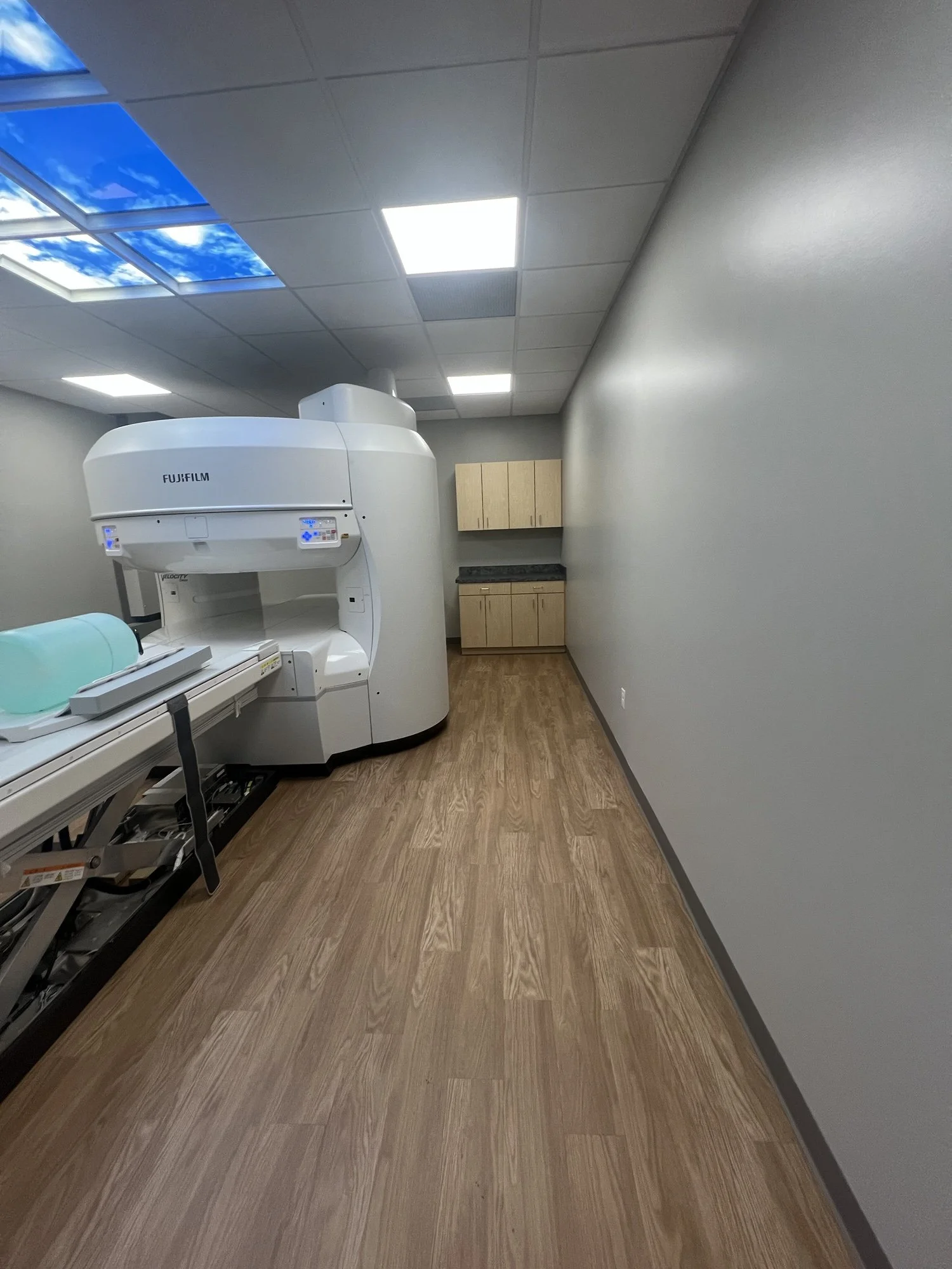 Medical imaging room with a large Fujifilm MRI machine near the wall, wooden cabinets in the background, wood flooring, and bright ceiling lights.