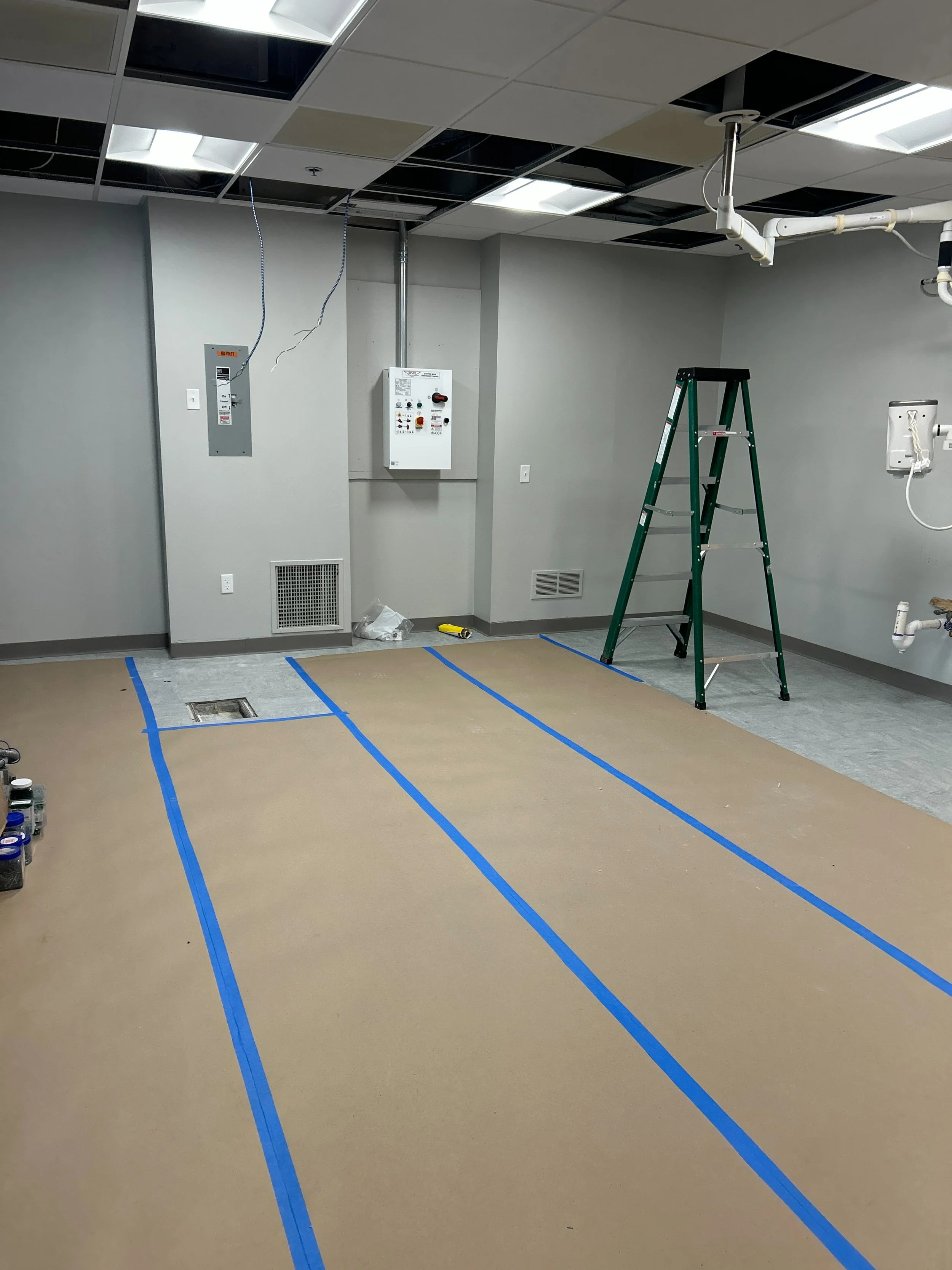 Empty room under renovation with electrical wiring, a ladder, and blue painter's tape on the floor.