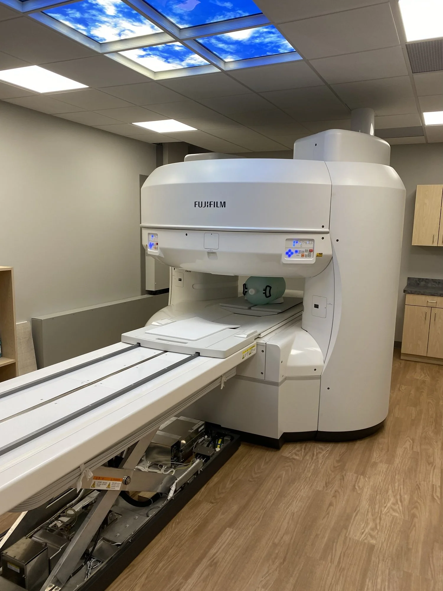 A medical MRI machine in a clinical room with a cartoon frog figure on the table inside the machine and a ceiling panel depicting a blue sky with clouds.