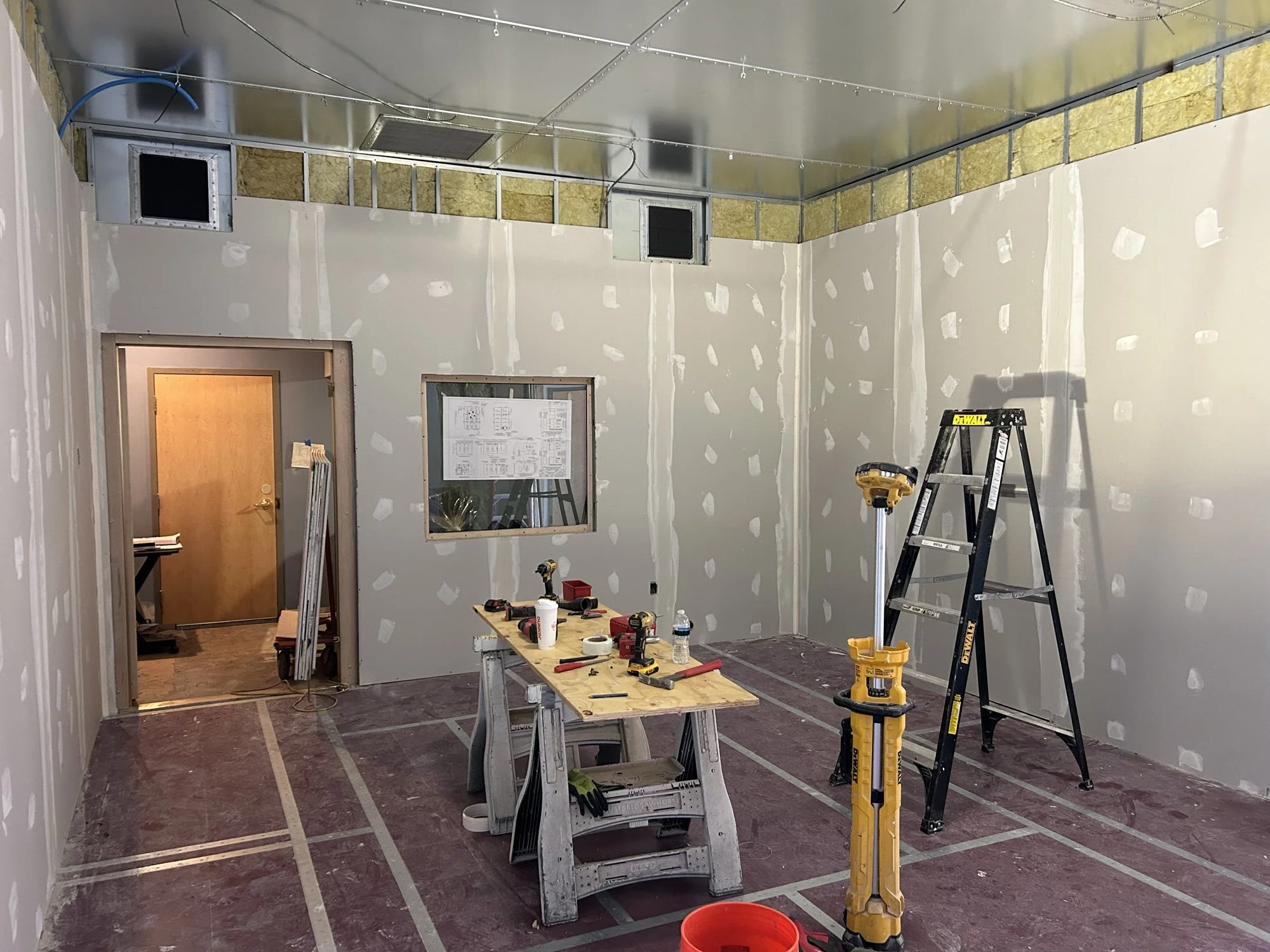 Room under construction with drywall in progress, tools on a plywood workbench, a ladder, a cordless drill, and a yellow pile driver, with taped markings on the floor and exposed ventilation ducts near the ceiling.