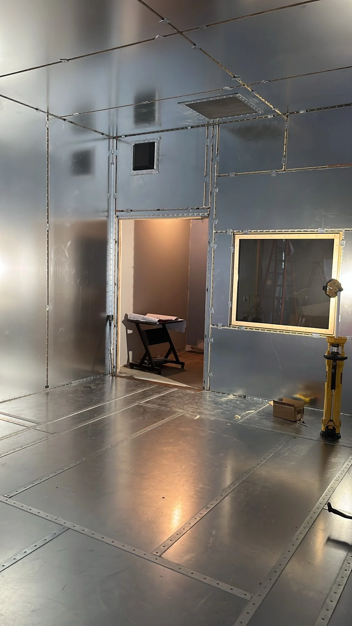 A photo of an unfinished room with metallic insulated walls and ceiling, with window and door openings. Construction tools and a workbench are visible.