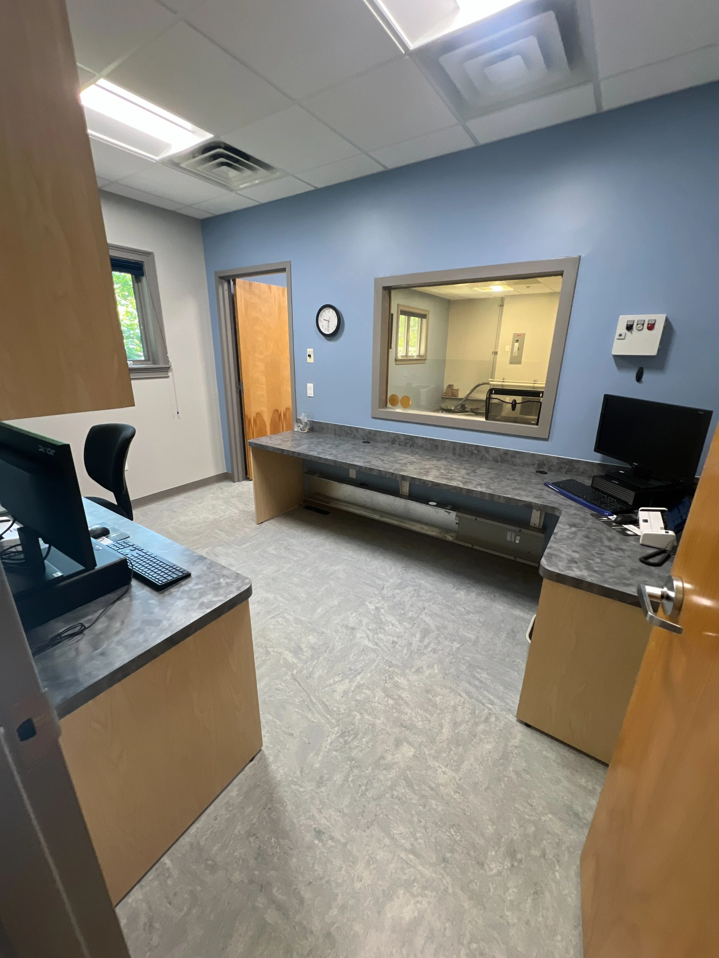 Empty medical office room with a counter, a computer, a window, and a door leading to an examination room.