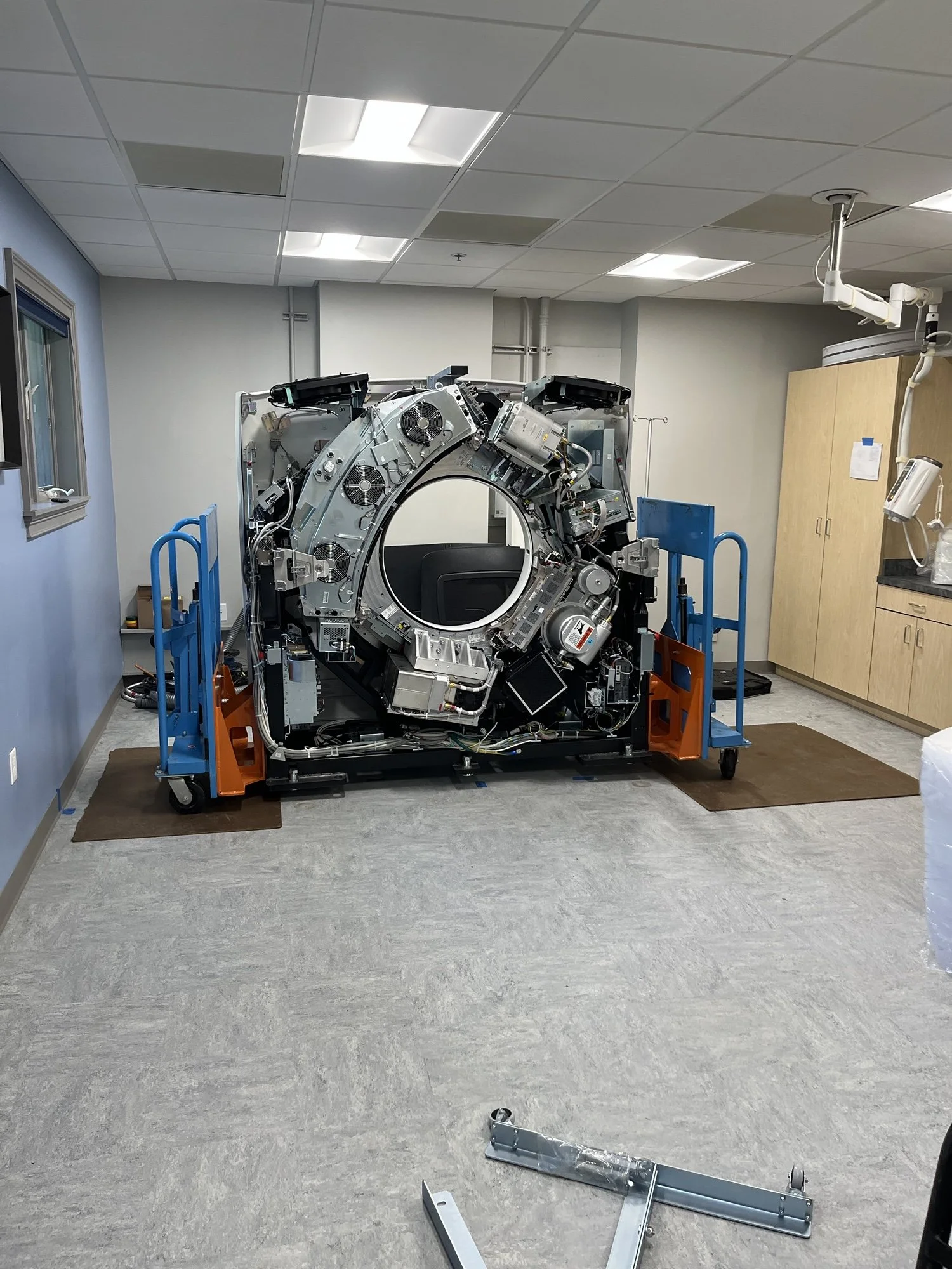 A large medical imaging machine, likely a CT scanner, in a room with beige walls, a window, cabinets, and medical equipment. The machine is partially disassembled or in maintenance, sitting on a blue and orange platform.