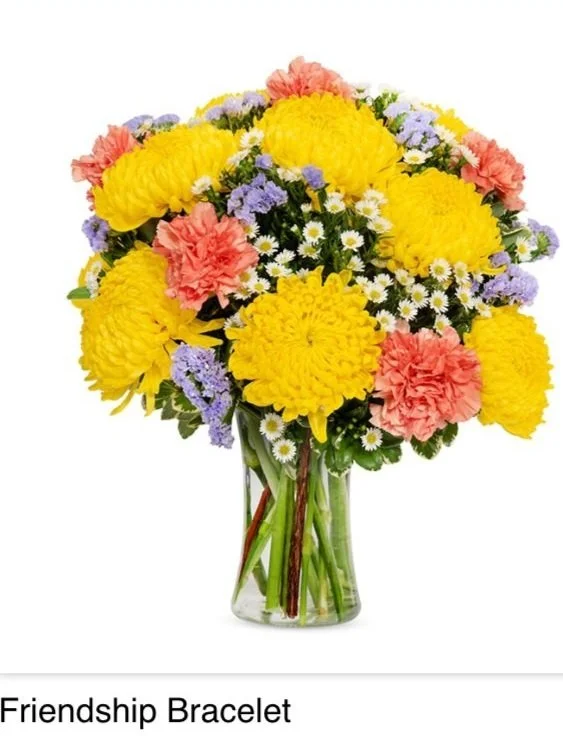 A bouquet of yellow, pink, purple, and white flowers in a clear glass vase, labeled as Friendship Bracelet.