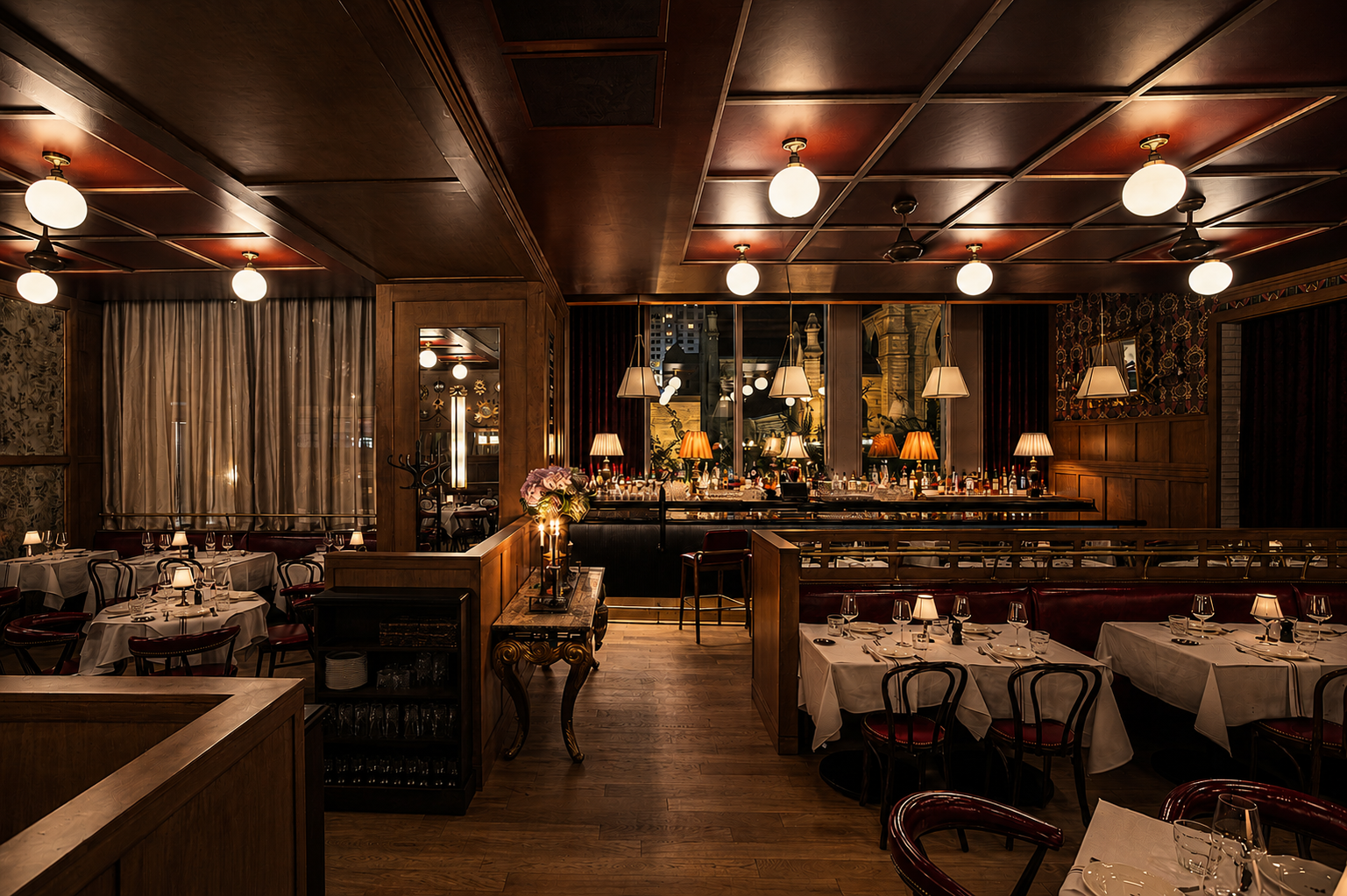 Elegant restaurant dining room with round tables covered in white tablecloths, set with glasses, napkins, and candles. The background features a bold red wall with mounted lamps and decorative branches, illuminated by unique modern chandeliers.