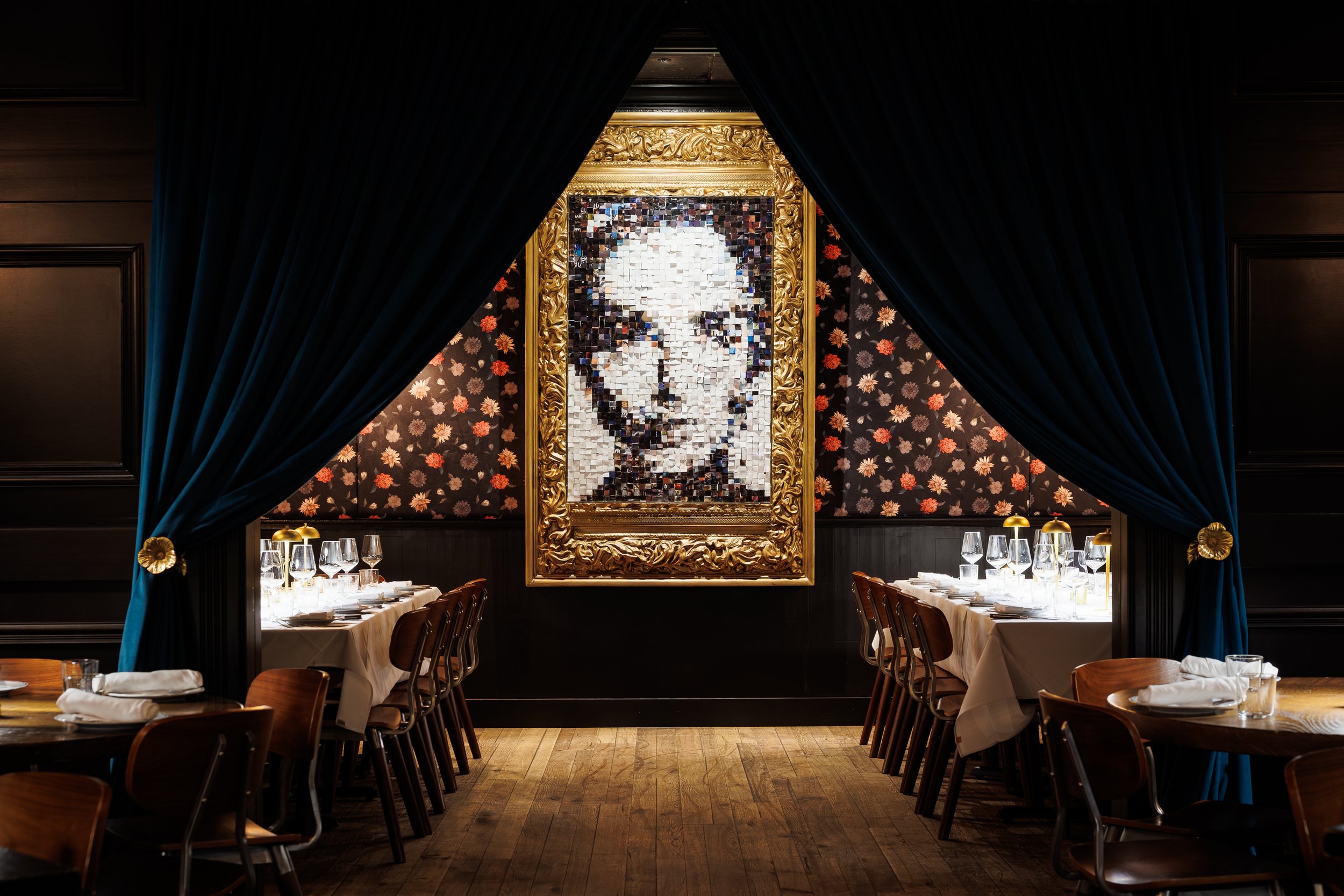 An elegant bar area with a circular bar counter, barstools, backlit liquor bottles, decorative flowers on the wall, a checkered floor beneath the bar, and a dimly lit dining table set with candles and glasses.