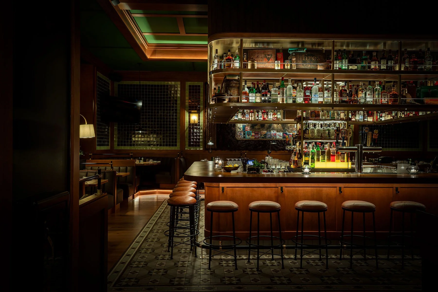 Empty bar with a wooden counter, high stools, and illuminated shelves stocked with liquor bottles in a dimly lit, stylish lounge or bar.
