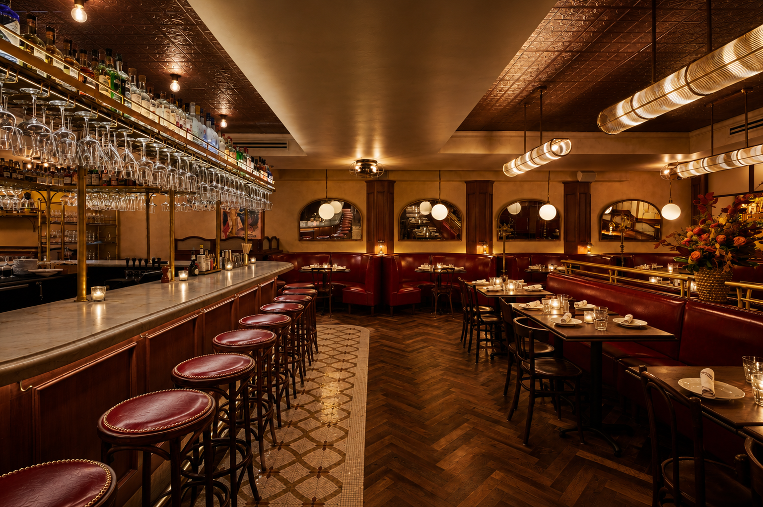 A cozy, dimly-lit bar with a central marble counter, surrounded by high wooden stools, ornate chandeliers, and a wall filled with liquor bottles and hanging glassware.