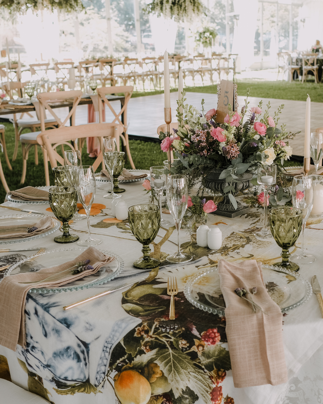 Wedding reception table decorated with a floral centerpiece, candles, glassware, plates, and napkins, set outdoors with chairs and a dance floor in the background.